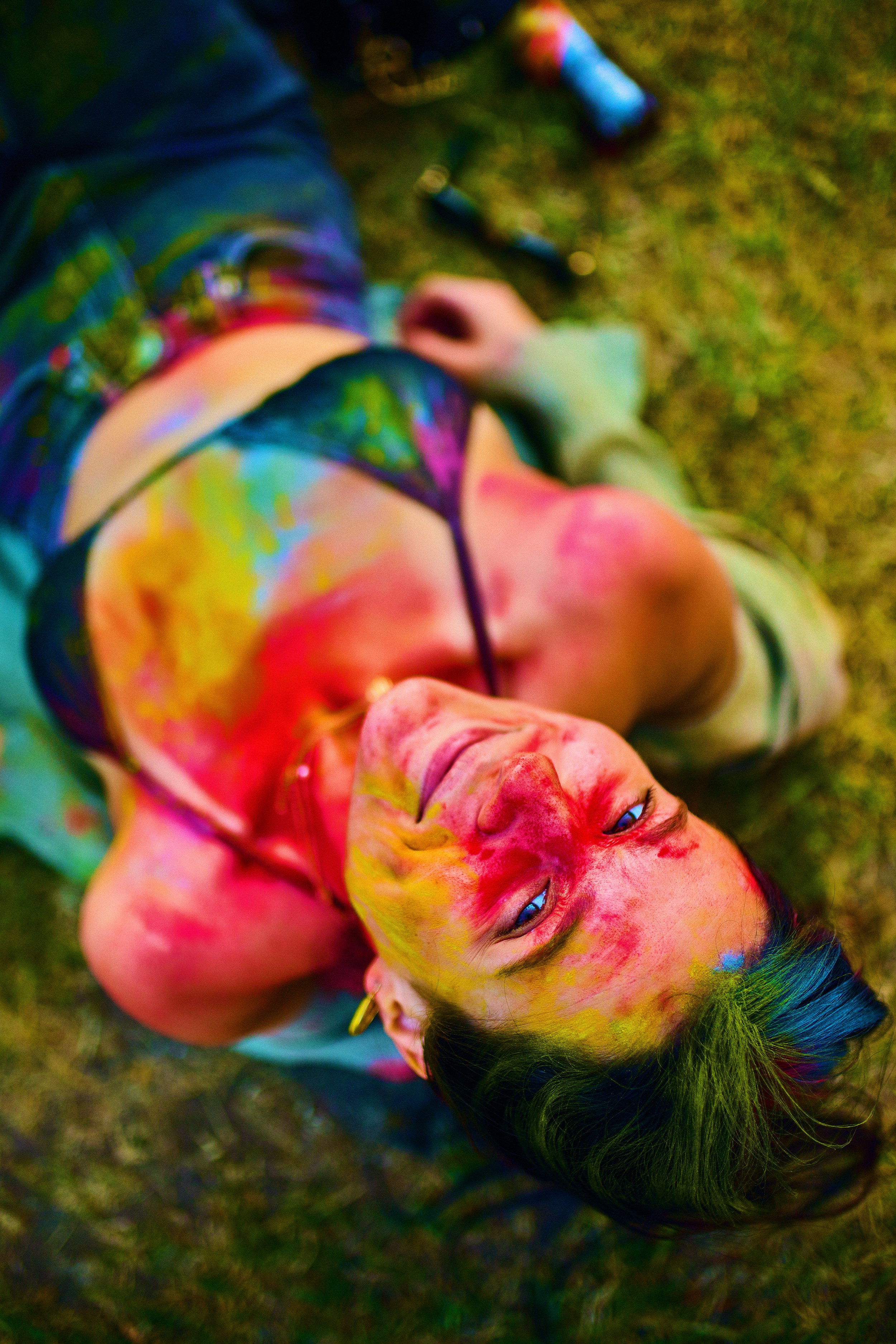 A woman lying on the grass covered in colorful paint splatters, looking up with a relaxed expression.