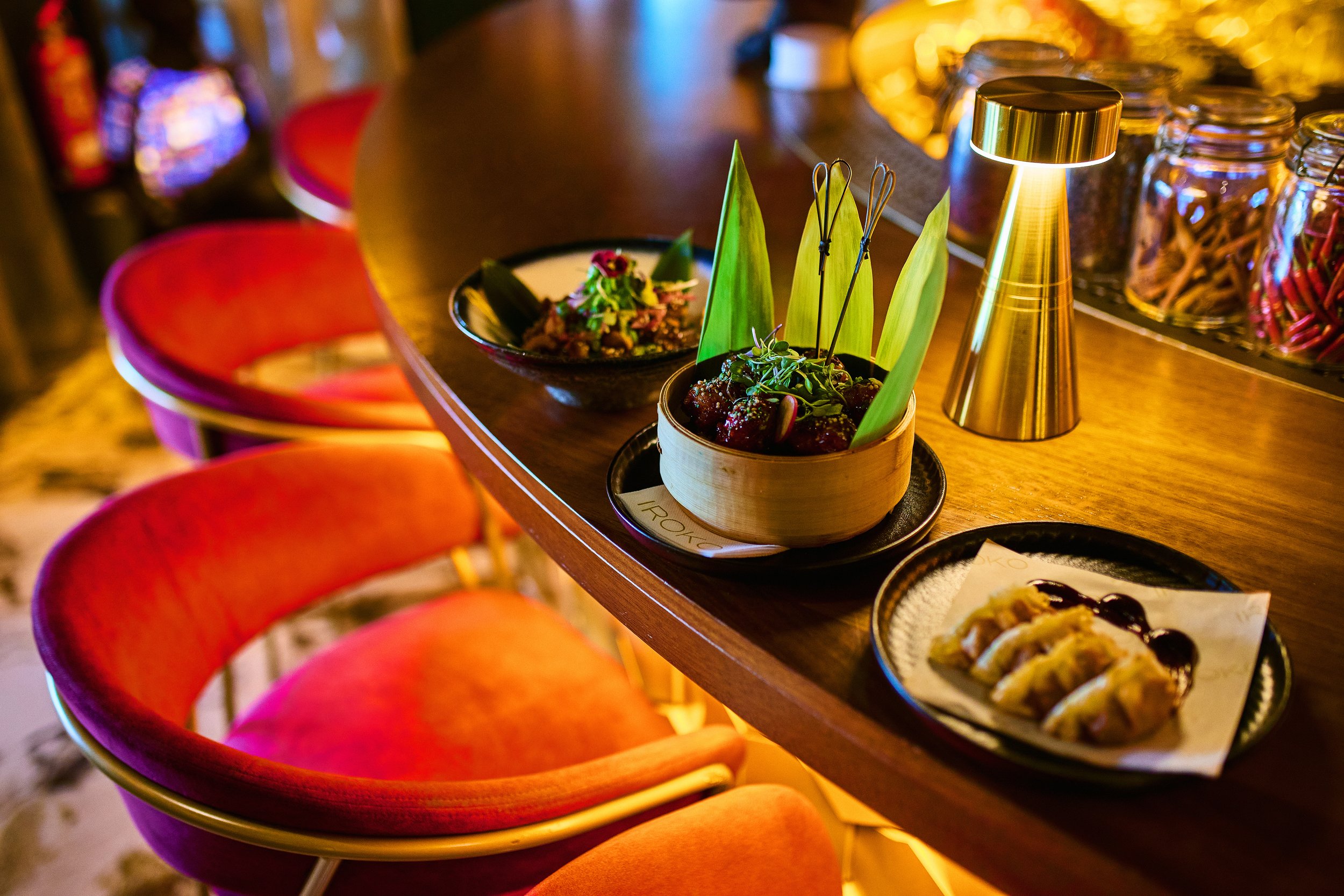 A restaurant bar counter with small plates of food, including a bowl of meatballs with garnishes, a rolled appetizer with sauce, and another dish with colorful ingredients, all illuminated by warm lighting, with red bar stools arranged along the coun