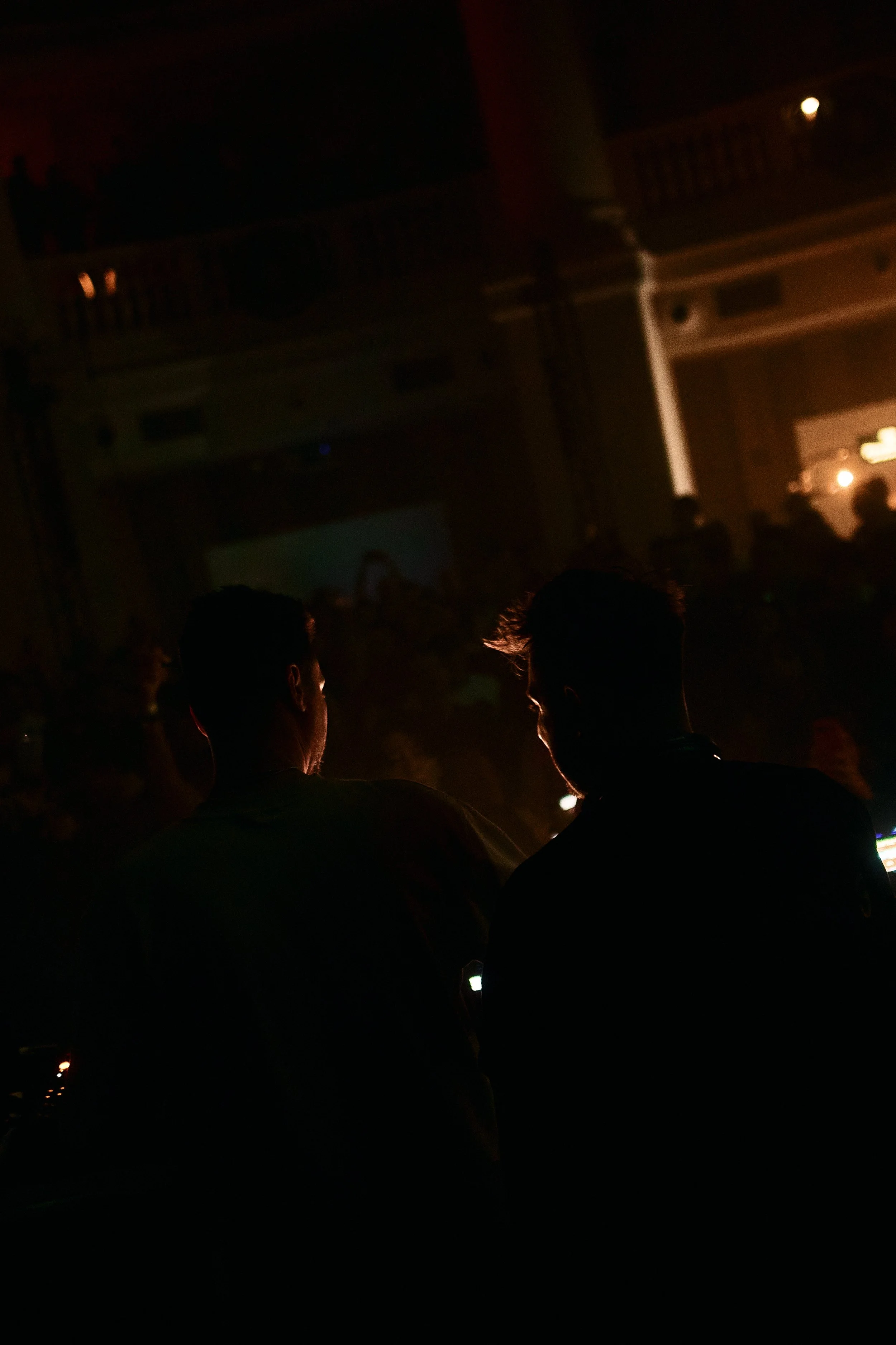 Silhouettes of two people in a dark, crowded room, possibly a nightclub or concert, with stage lights and a balcony in the background.