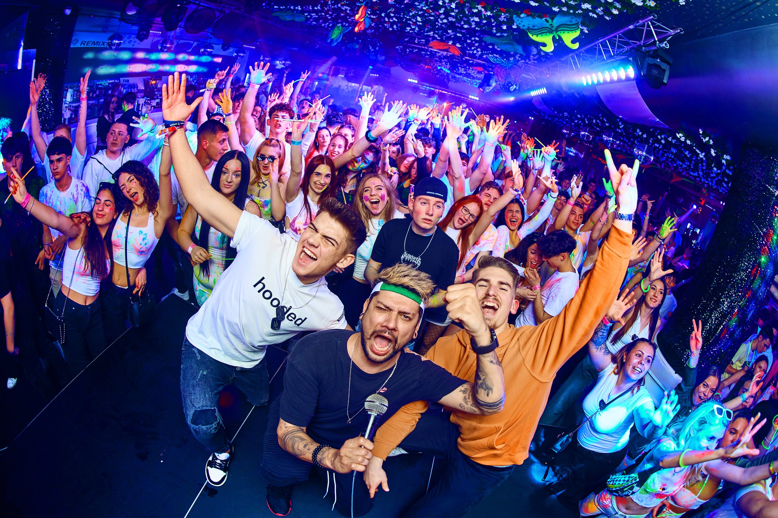 A large group of young people at a party with colorful lighting, many raising their hands and smiling, some making peace signs, with three people in front posing enthusiastically with one holding a microphone.