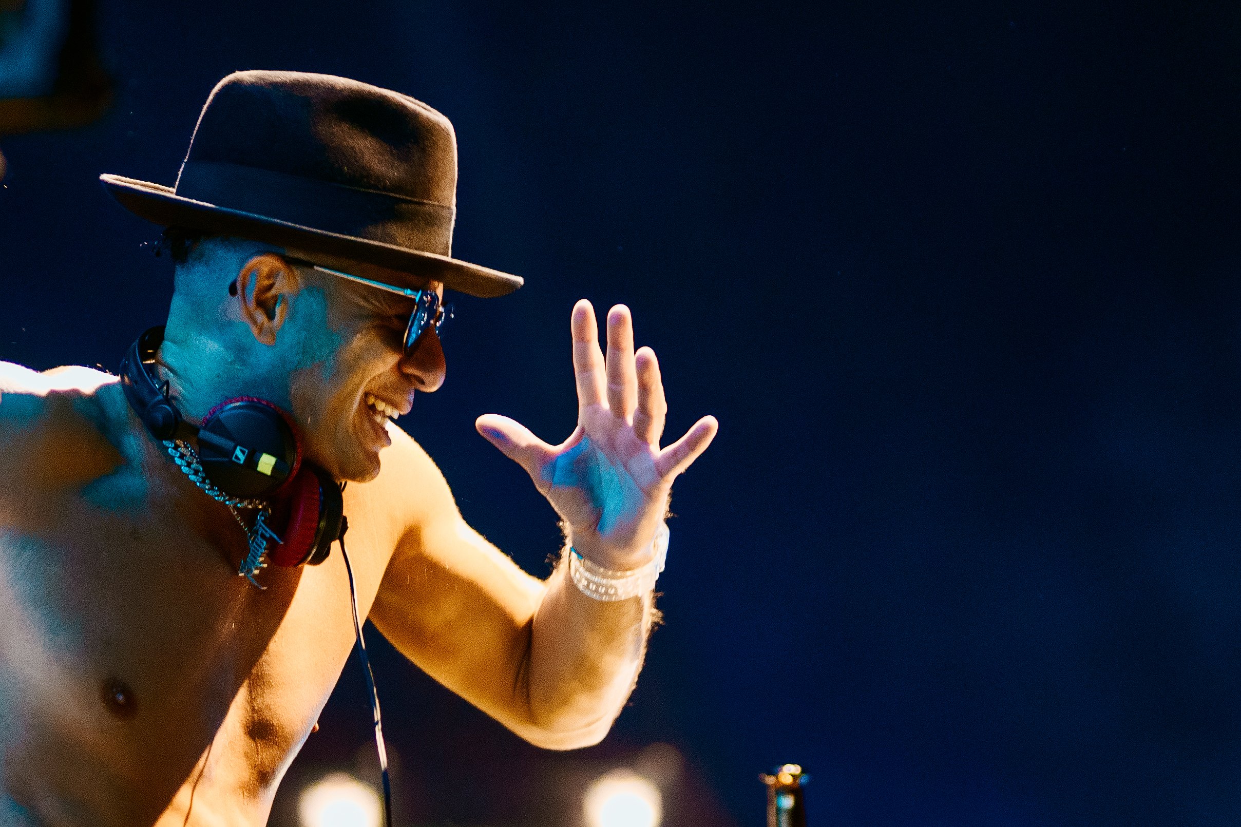A shirtless man wearing sunglasses, a hat, and a bracelet, smiling and waving at night against a dark sky, with DJ headphones around his neck.