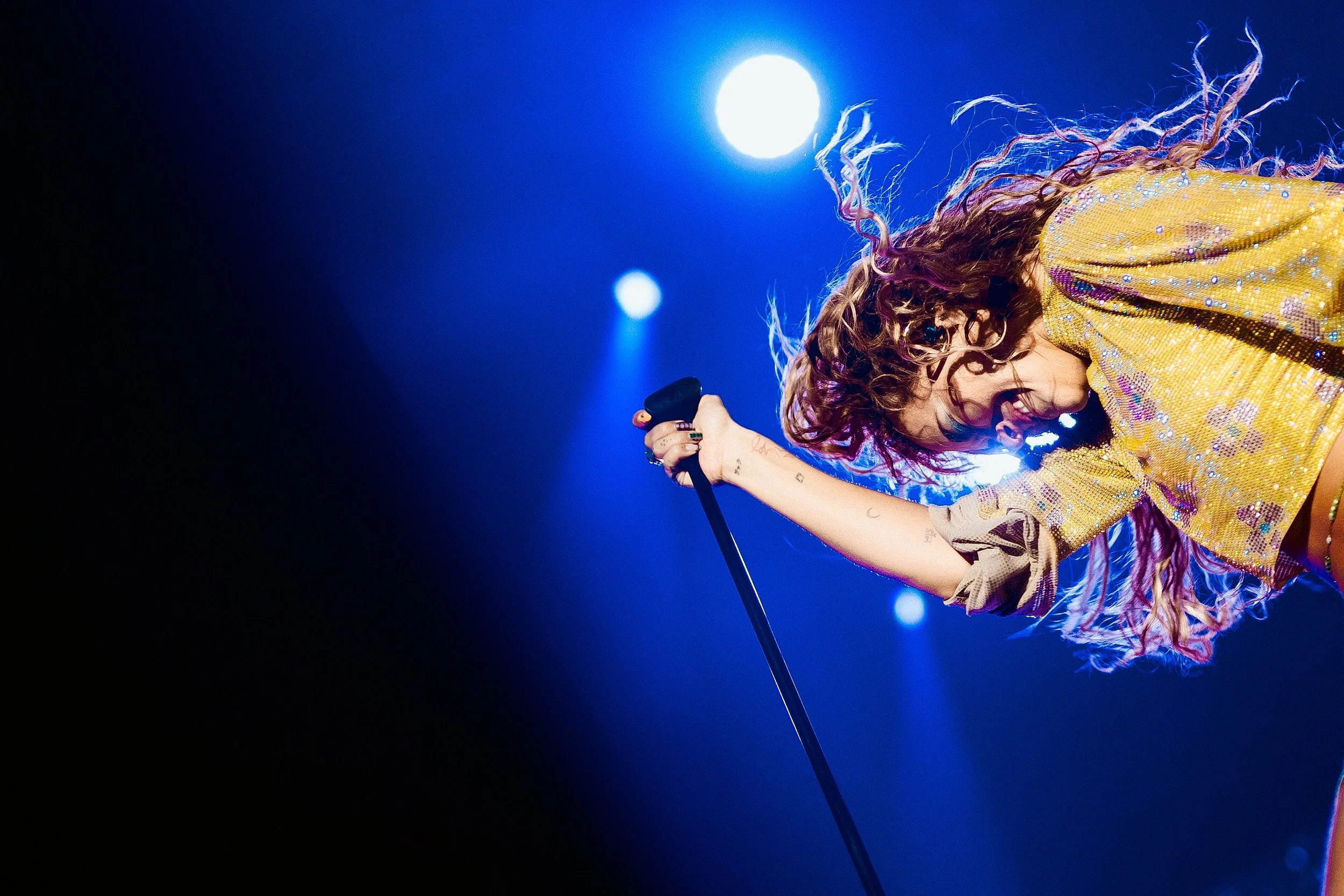 A female singer with curly hair singing passionately on stage, illuminated by bright blue and white stage lights, wearing a yellow sequined top.