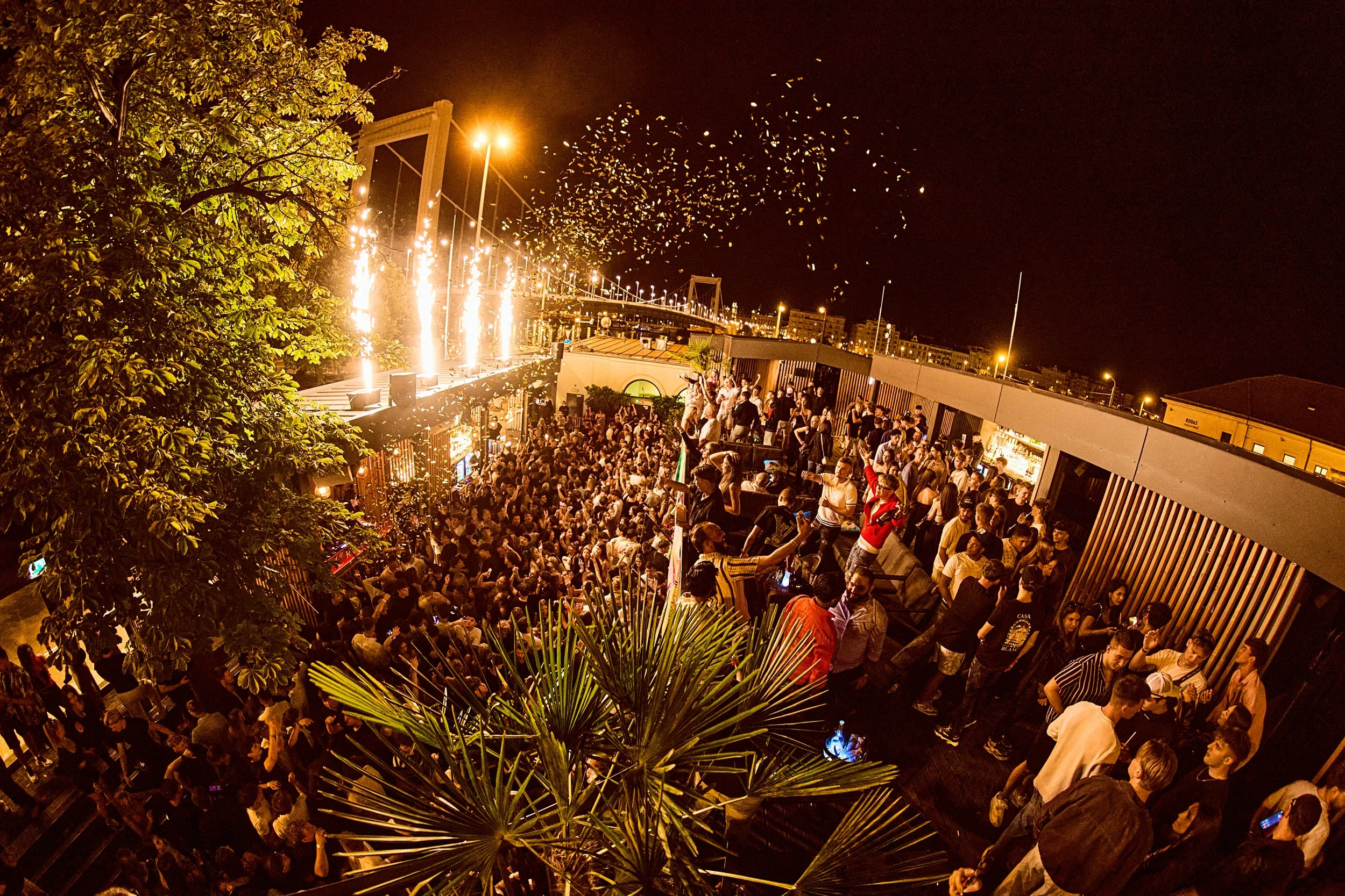 Nighttime rooftop party with many people dancing and mingling, fireworks, and sparklers in the sky, with city buildings in the background.