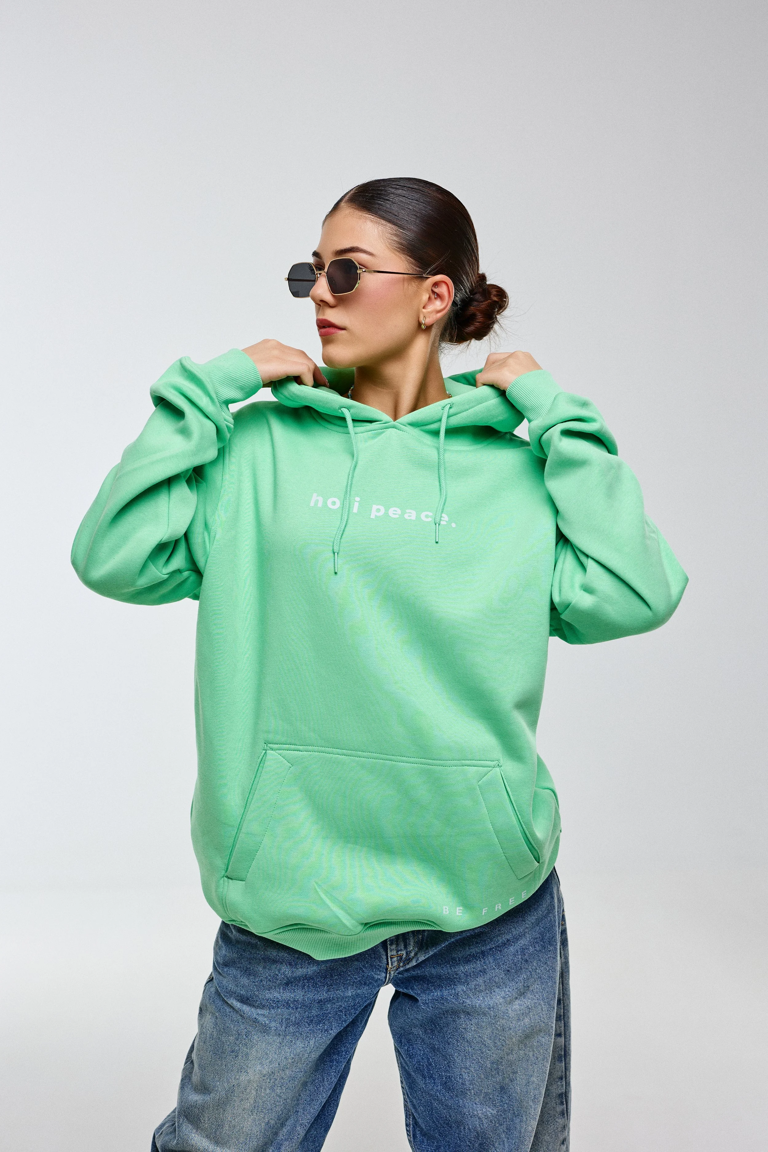 A woman wearing a mint green hoodie with text on it, black sunglasses, and blue jeans, adjusting her hoodie against a plain background.