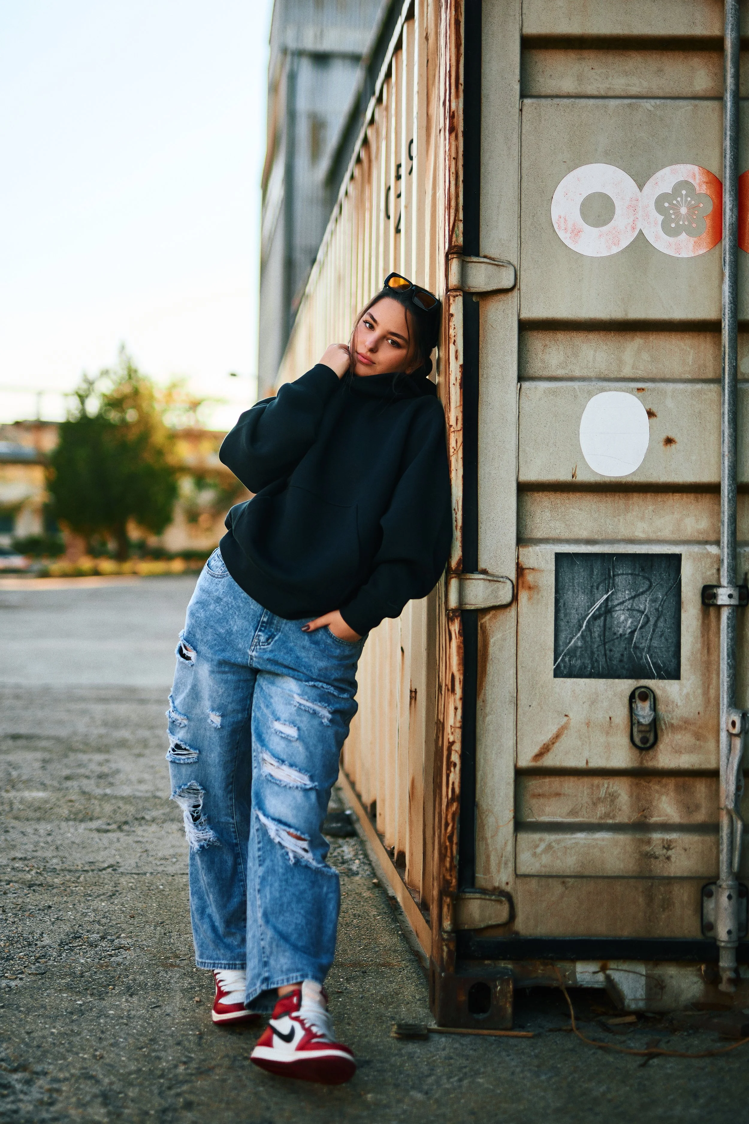 Young woman leaning against a rusty shipping container outdoors, wearing a black hoodie, ripped jeans, and red and white sneakers.