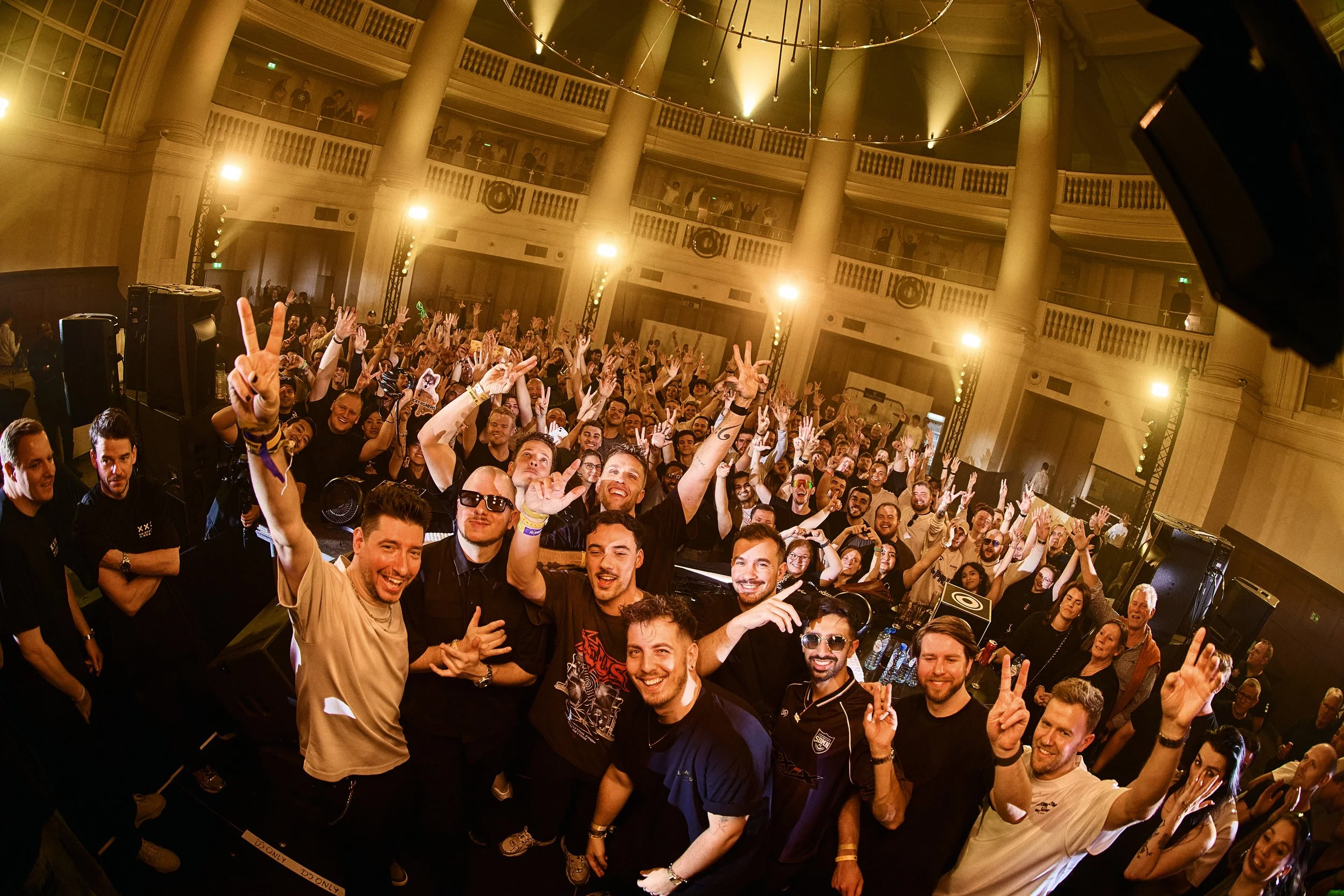 A large crowd of people at an indoor concert or party, with some on a stage or DJ booth in the foreground, many smiling, raising their hands, and making peace signs or other gestures. The venue has tall columns and a high, rounded ceiling with lighti