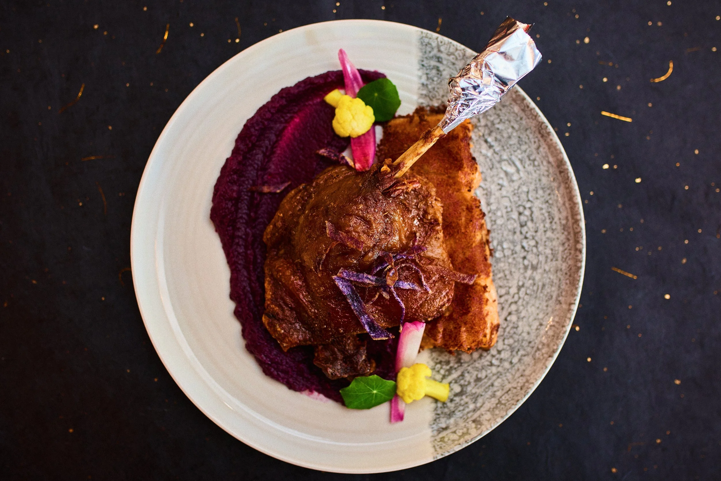 A plated gourmet dish featuring a piece of cooked meat with sauce, served with a purple puree and garnished with edible flowers and pickled vegetables on a white and grey plate.