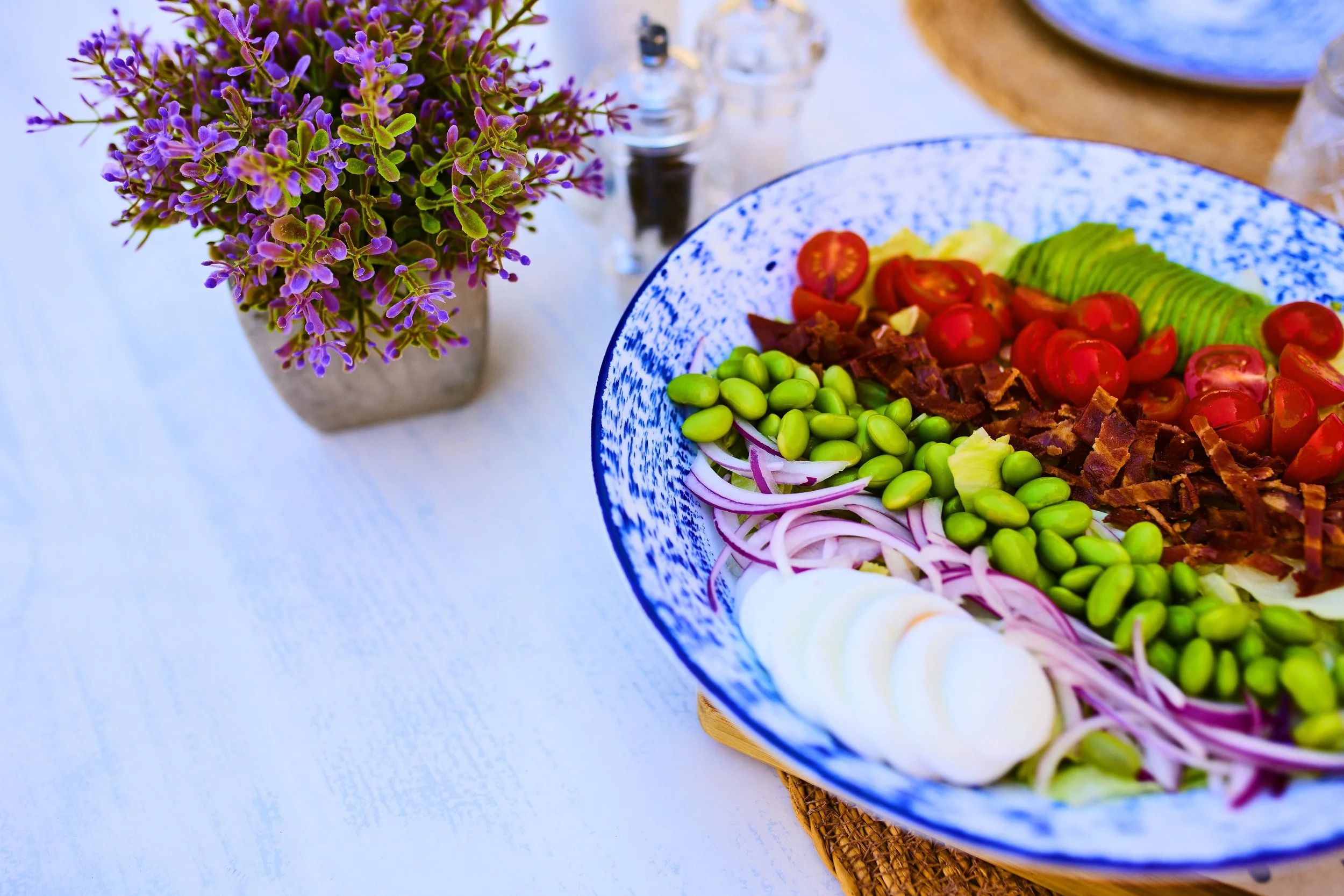 A colorful salad in a blue and white patterned bowl featuring cherry tomatoes, sliced avocado, chopped bacon, green beans, red onion, and sliced boiled eggs, with a purple flowering plant and salt and pepper shakers on a white table in the background
