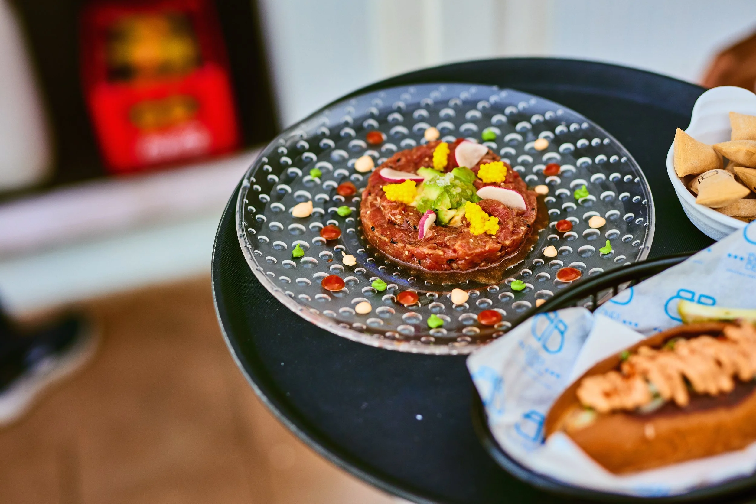 A dish of steak tartare garnished with vegetables and colorful toppings on a black plate, with other similar dishes partially visible.
