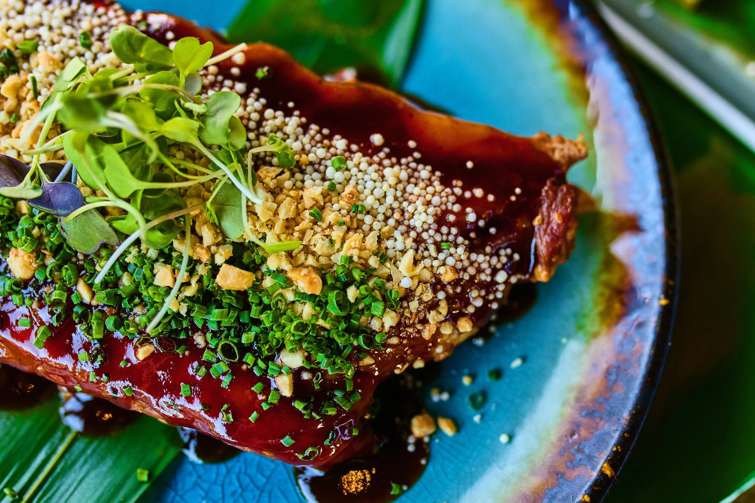 Close-up of a gourmet dish with garnished microgreens, crushed nuts, and small white spheres on a glazed blue-green ceramic plate.