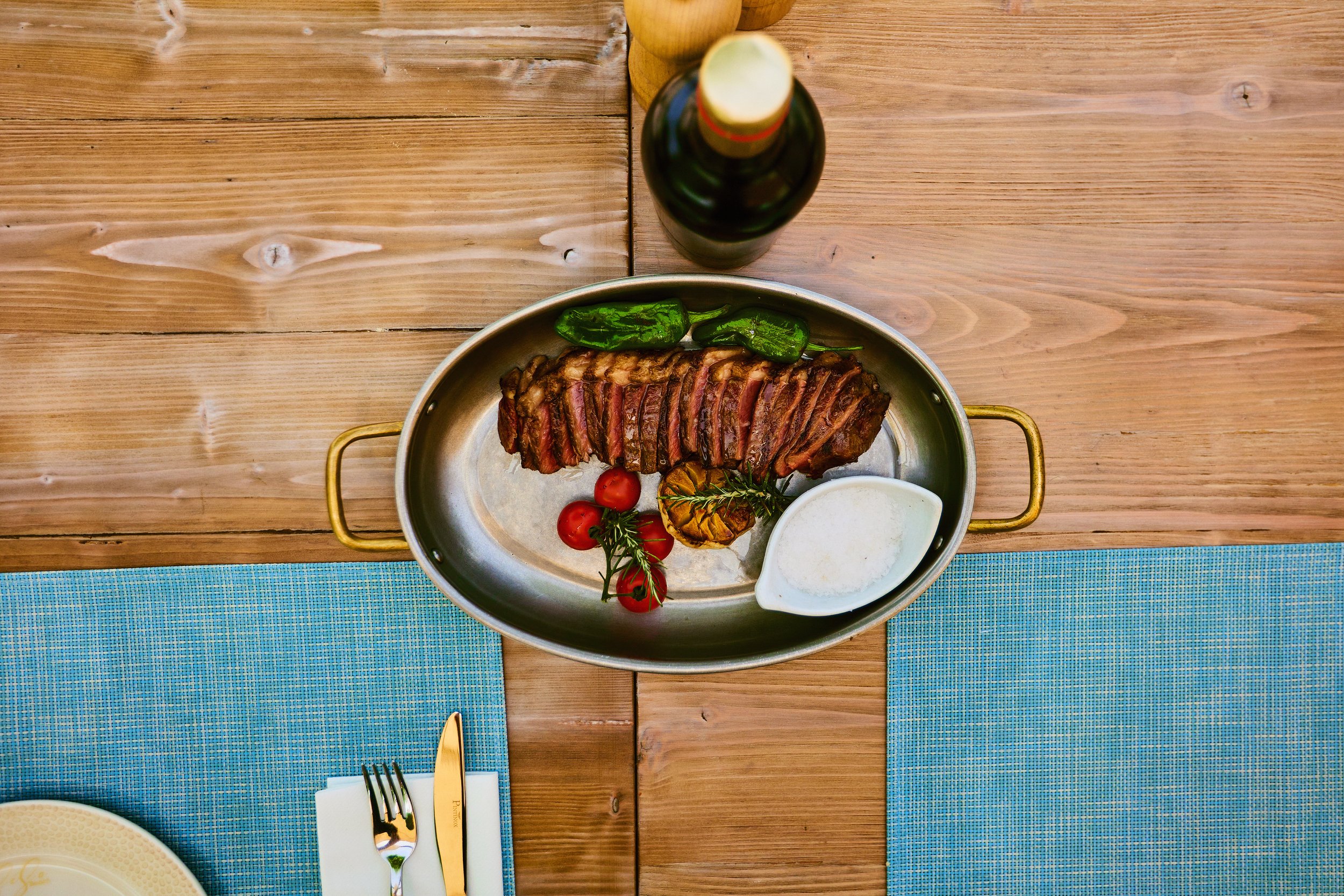 A grilled steak served with cherry tomatoes, green peppers, a roasted garlic bulb, and a white sauce in a white gravy boat on a metal platter with a bottle of wine in the background on a wooden table.