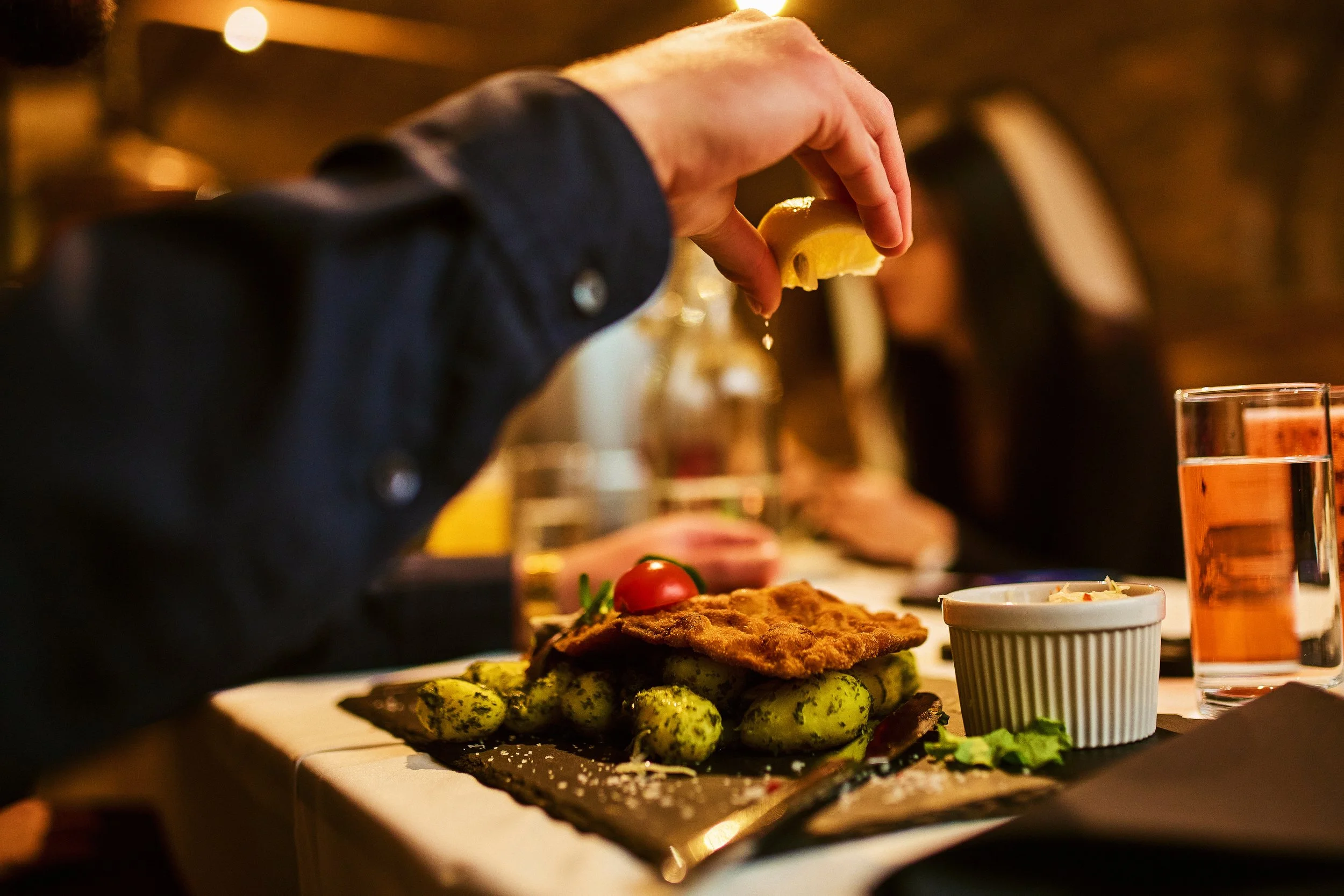 Person squeezing lemon over a plate of food including fried fish, cucumbers, and a cherry tomato, with a glass of rosé wine on the side in a dimly lit restaurant