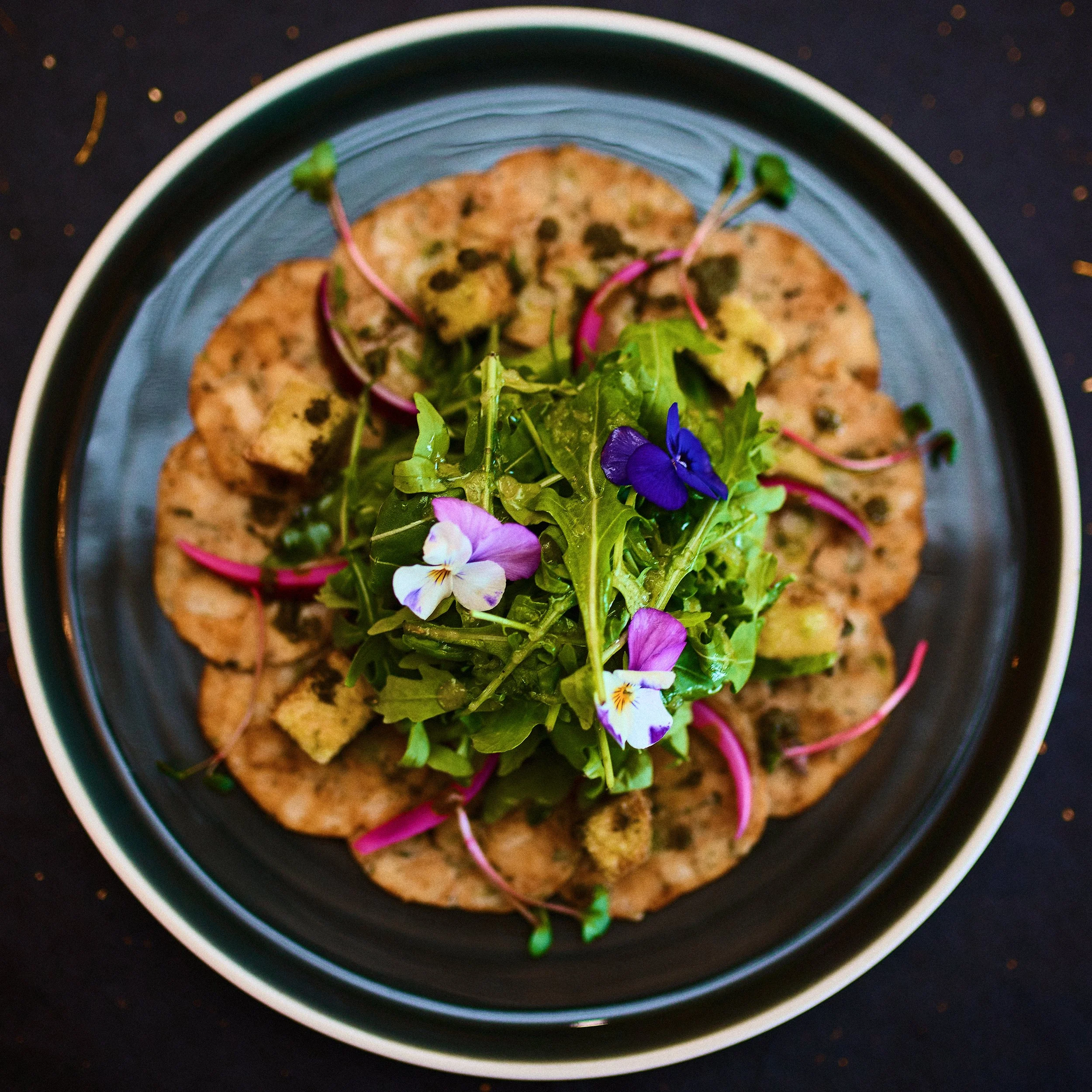 A black plate with a white rim, filled with a bed of mixed greens topped with colorful edible flowers, slices of radish, and croutons.