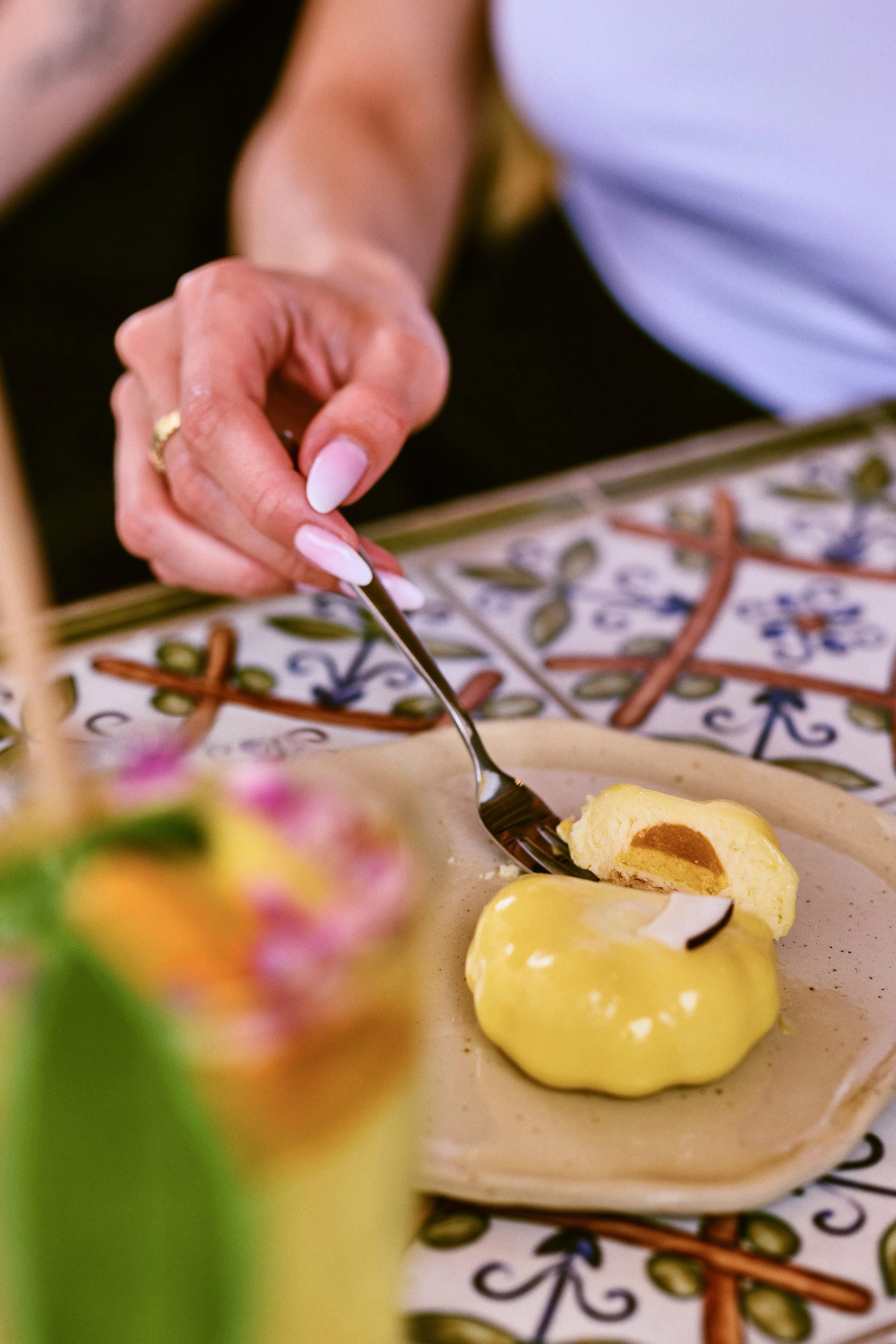 A person with manicured nails is cutting into a yellow dessert, possibly a cake or pastry, with a fork on a plate. The background features a decorative tiled table surface, and there is a blurred dessert or pastry in the foreground.