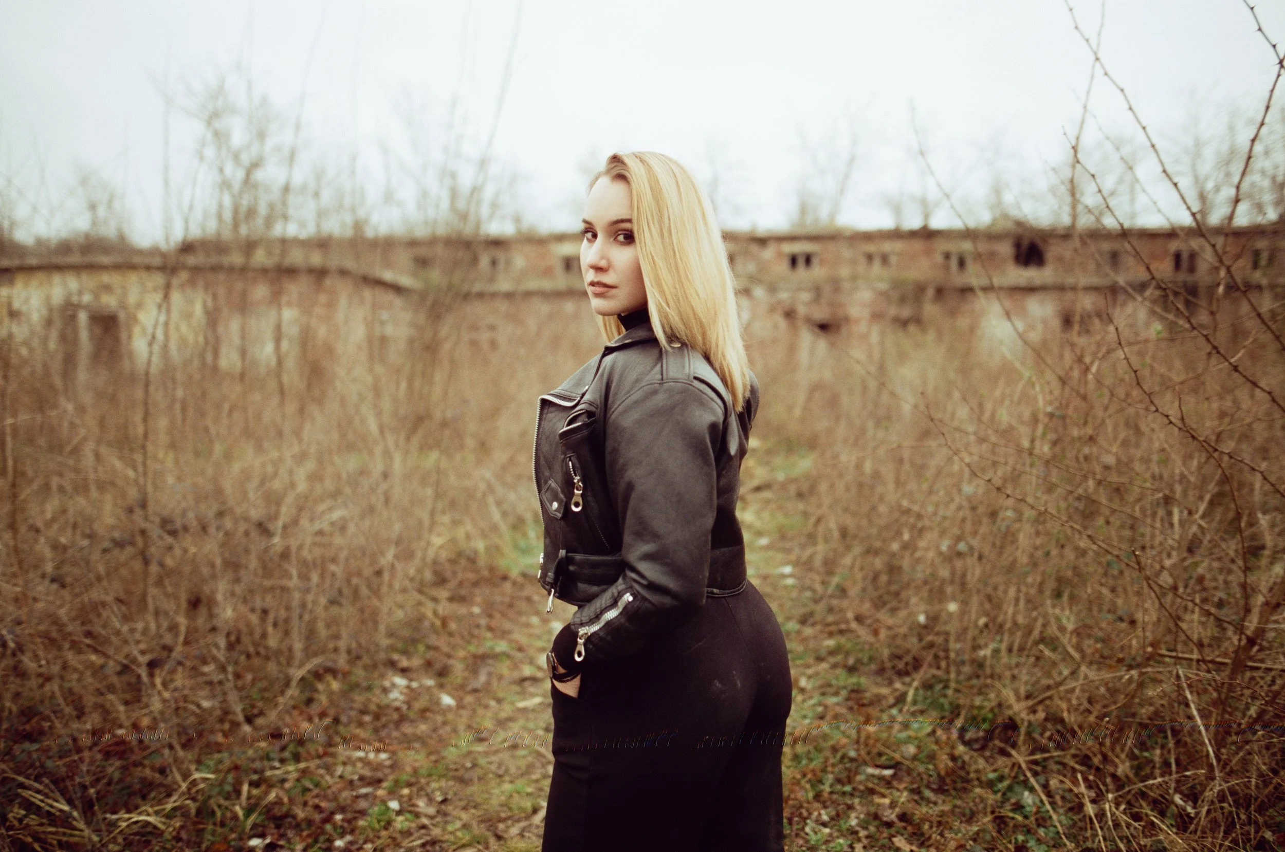 A young woman with blonde hair wearing a black leather jacket and black pants standing outdoors amid leafless bushes, looking towards the camera with an overcast sky in the background.