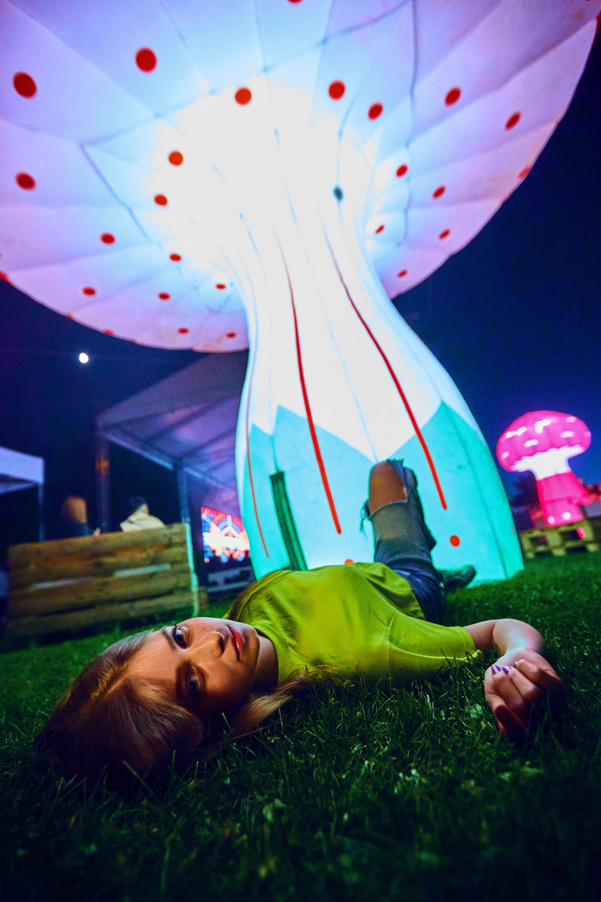 A woman lying on the grass at night under a large, illuminated mushroom-shaped display with red spots. There are other glowing mushroom installations visible in the background.