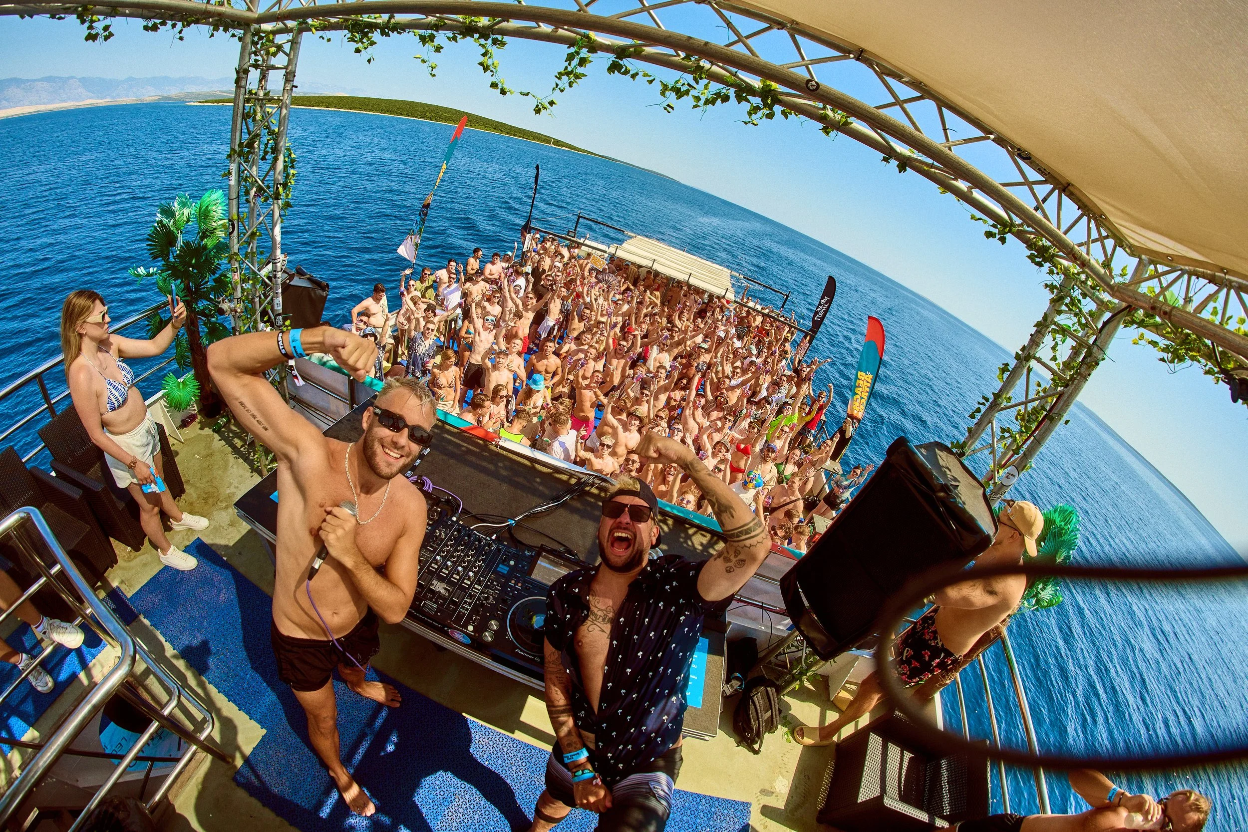A vibrant outdoor music event on a boat, featuring two shirtless DJs in sunglasses, with a large crowd of people dancing on the boat deck and the ocean in the background.