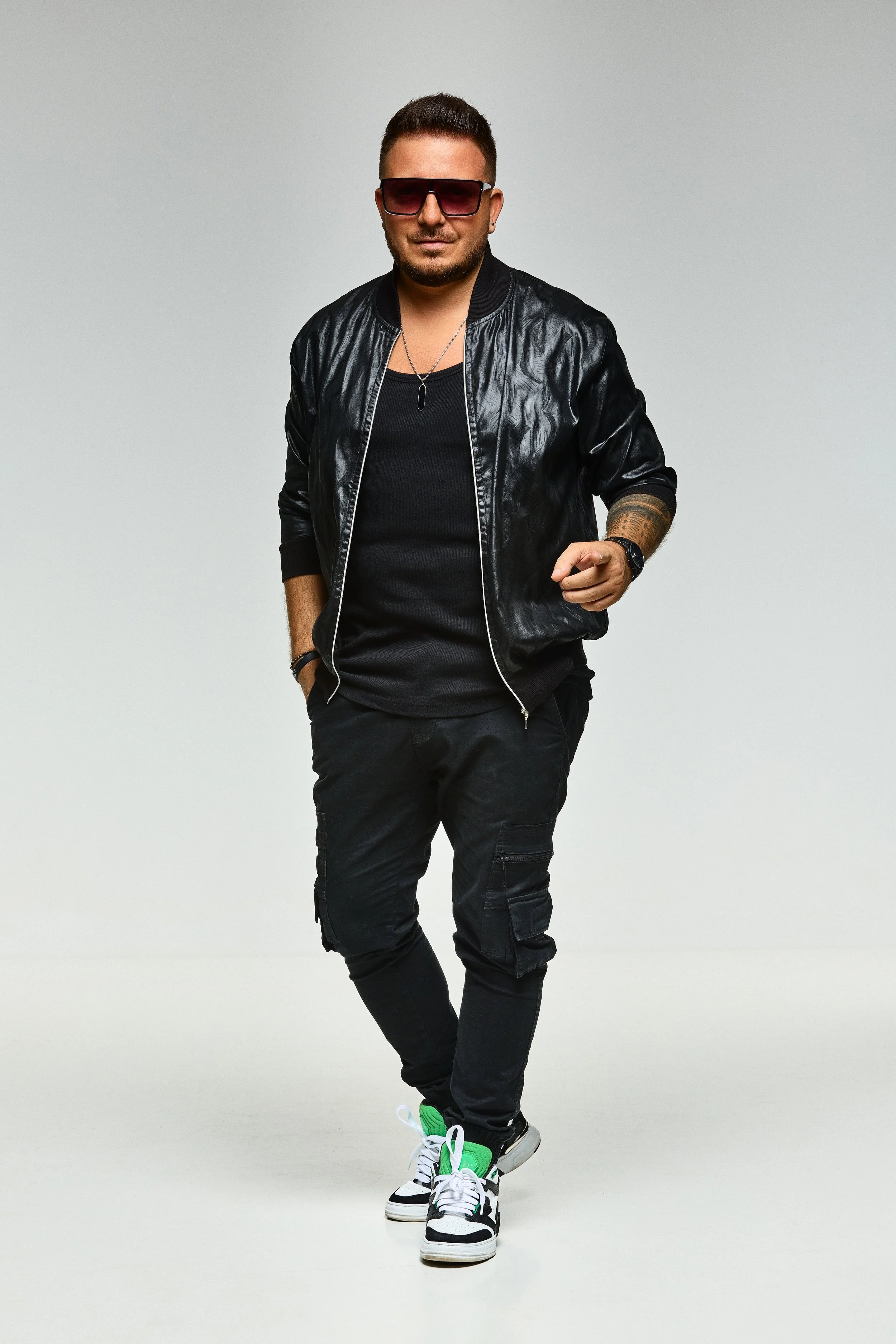 A man wearing sunglasses, a black leather jacket, black pants, and sneakers, walking confidently against a plain background.