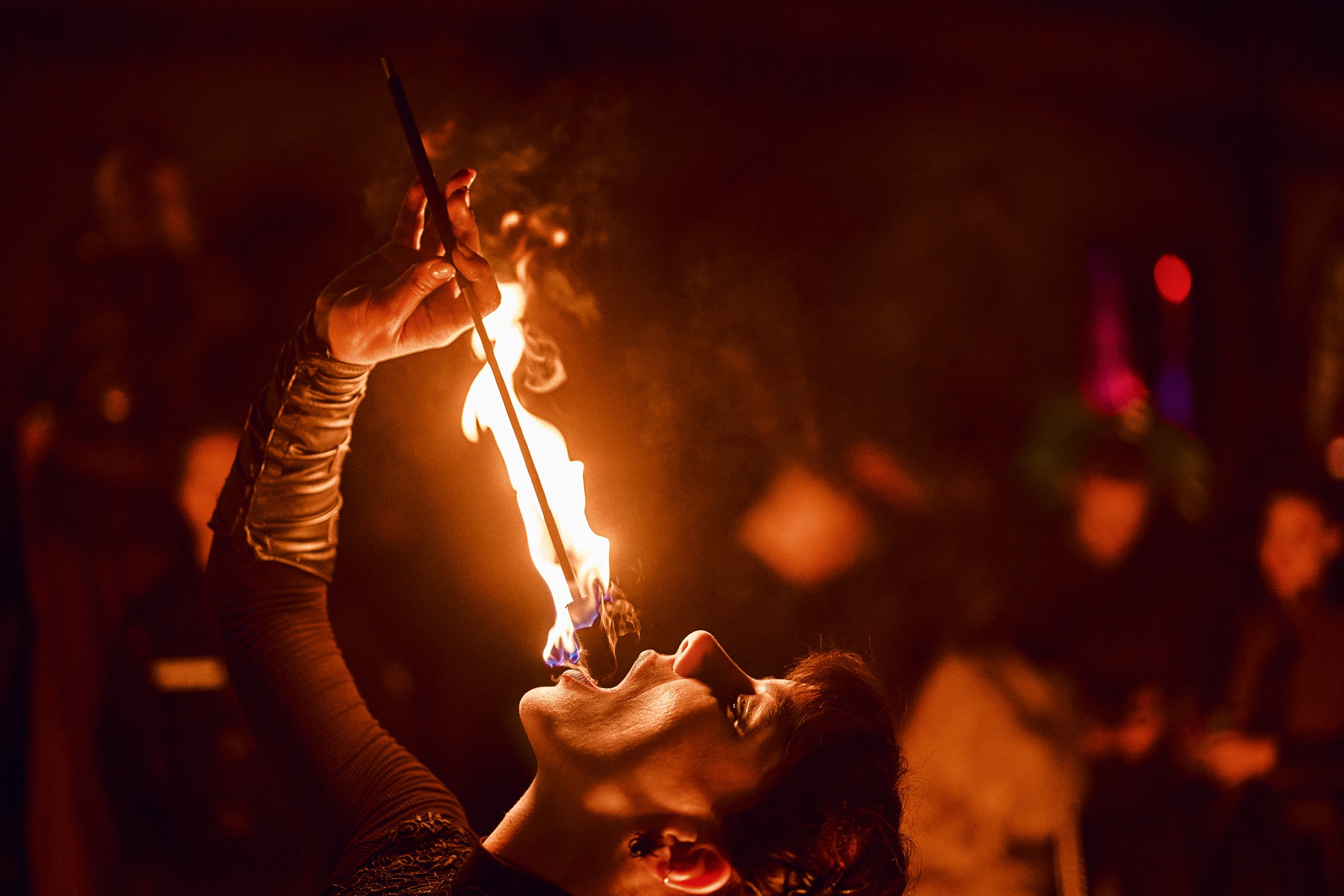 A woman performing a fire-eating act at night, holding a lit torch close to her open mouth with flames emerging, surrounded by a dark crowd.