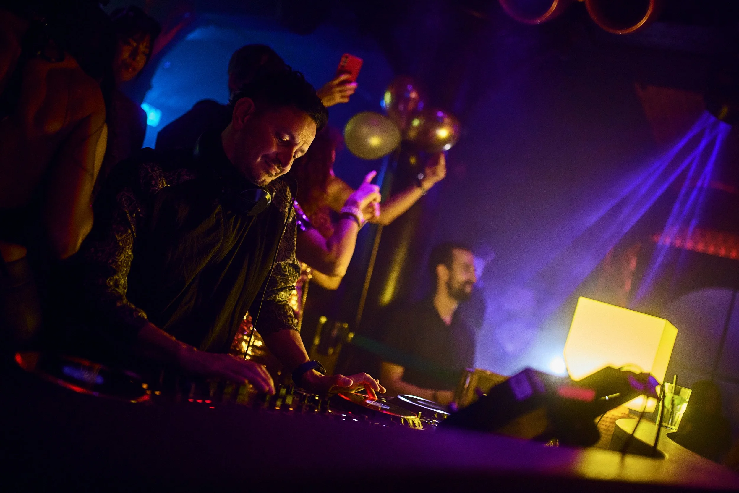 A DJ is performing at a nightclub with colorful lighting, surrounded by partygoers, some holding balloons and taking photos.
