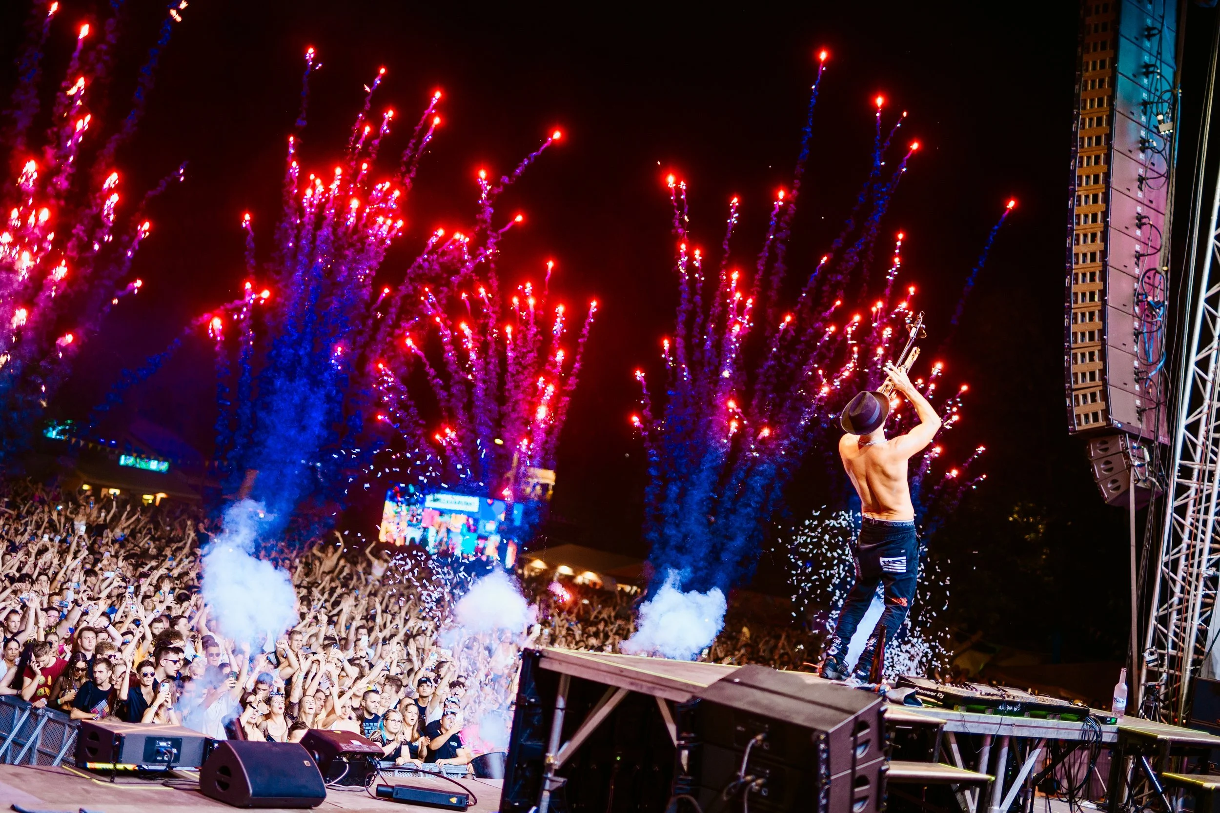 A performer on stage with a hat, facing away, holding a guitar above his head during a fireworks display at a concert.