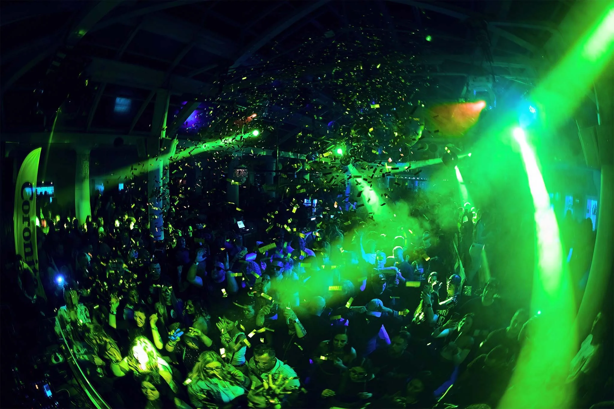 Crowd of people dancing and socializing in a nightclub with green and blue lighting, confetti, and laser effects.