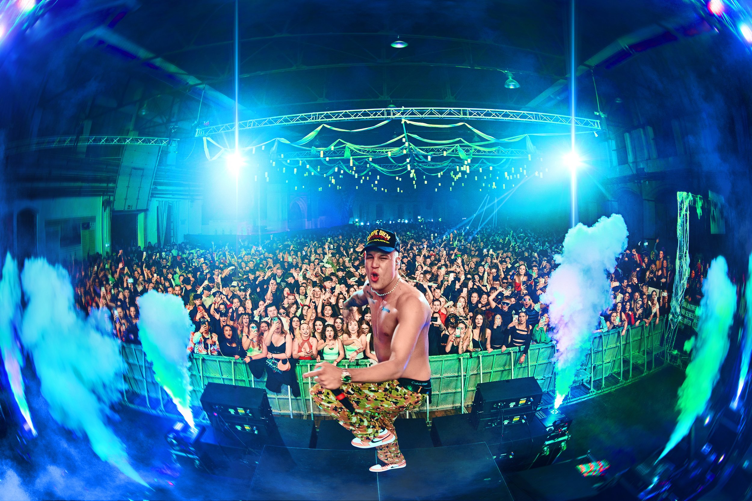 A shirtless performer in camouflage pants and a cap, holding a microphone, is on stage in front of a crowded crowd at a concert or music festival, with colorful smoke effects and blue lighting.