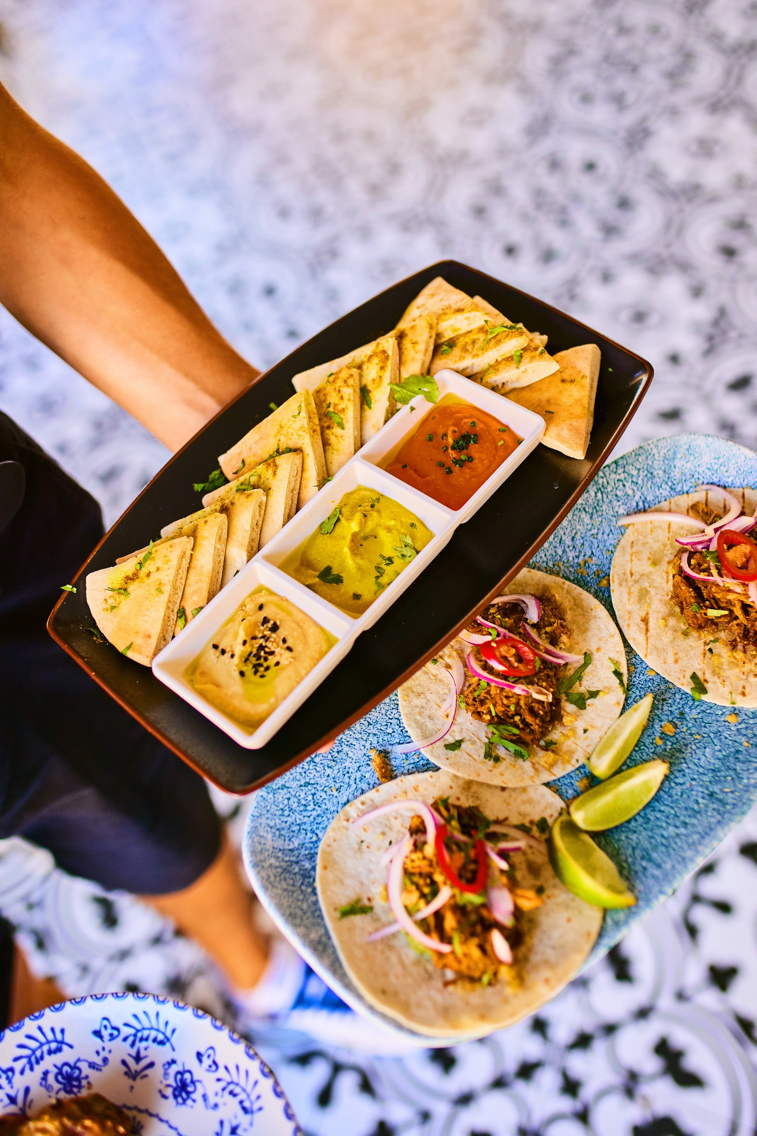 A hand holding a rectangular black plate with grilled quesadillas and three small bowls of dipping sauces; a side of tacos with shredded meat, onions, and peppers with lime wedges on a blue plate.