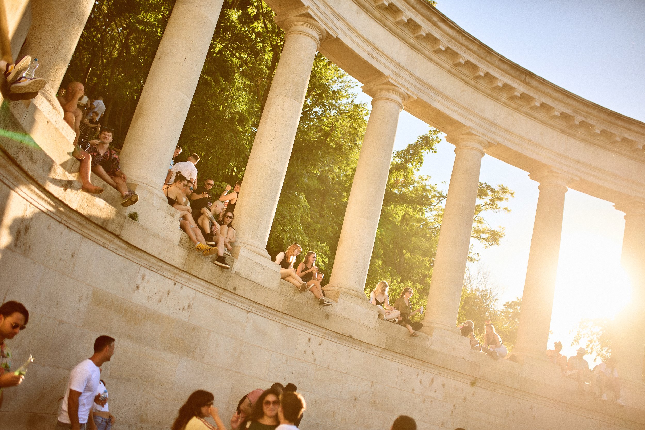 People sitting and standing on the steps of a large, classical stone structure with tall columns, illuminated by sunlight, surrounded by trees.