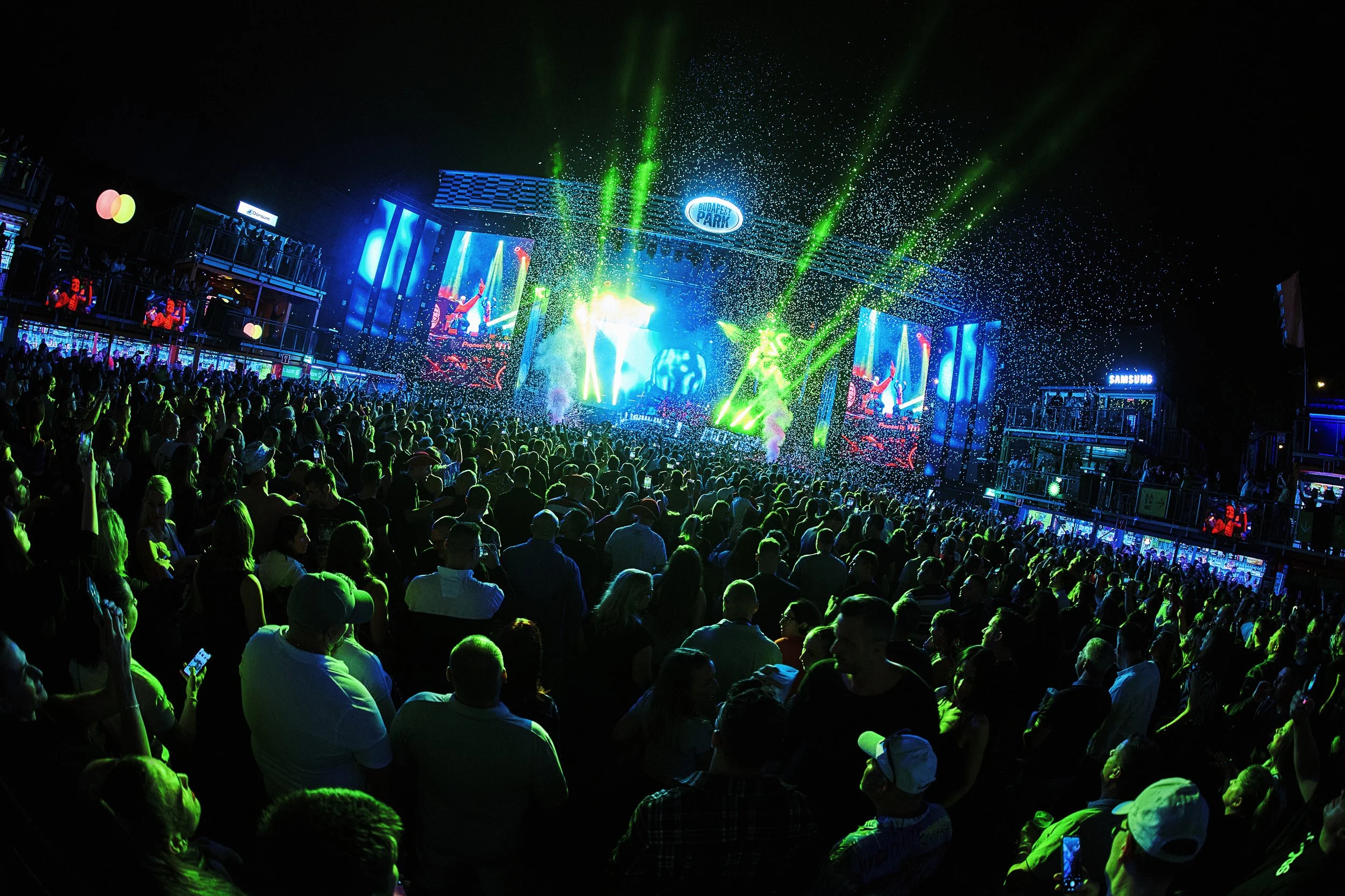 A large crowd at an outdoor concert at night with colorful stage lights, screens, and confetti in the air.