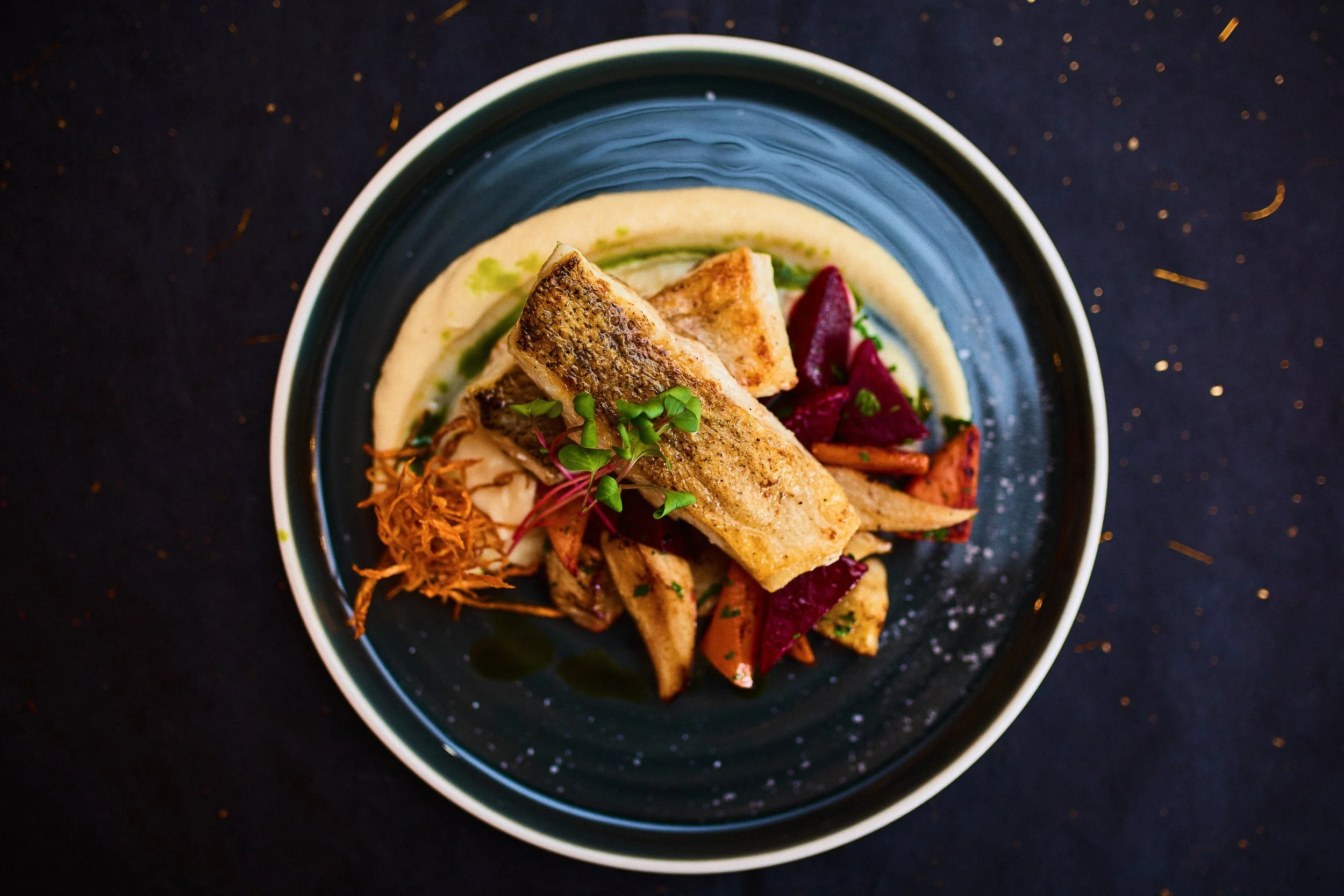 Pan-seared fish fillet served over roasted vegetables on a black plate with sauce and microgreens garnish.