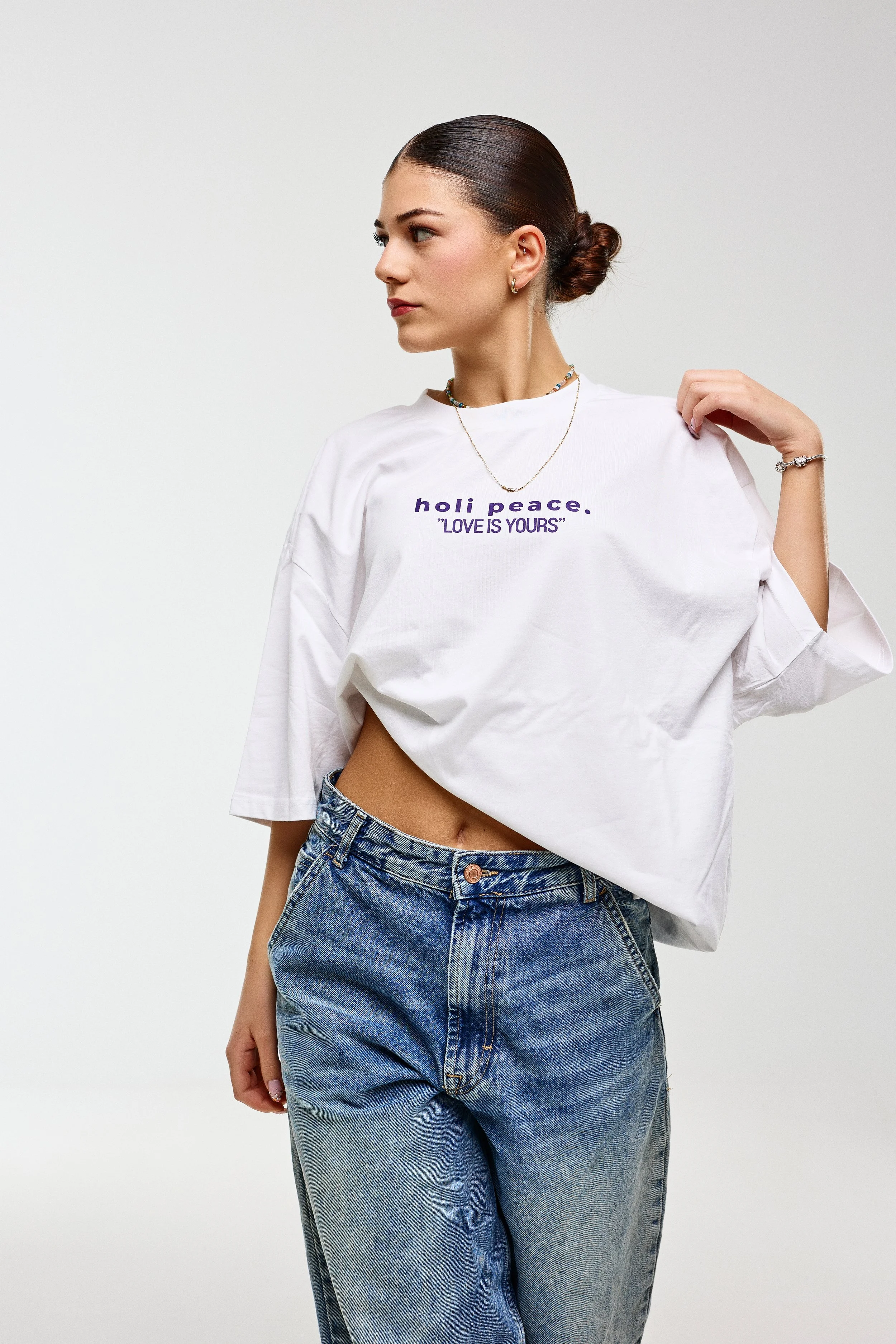 Young woman with dark hair tied back, wearing a white T-shirt with purple text that reads 'holi peace. 'LOVE IS YOURS',' and blue jeans. She has jewelry including earrings, a necklace, and a bracelet. She is standing against a plain white background.