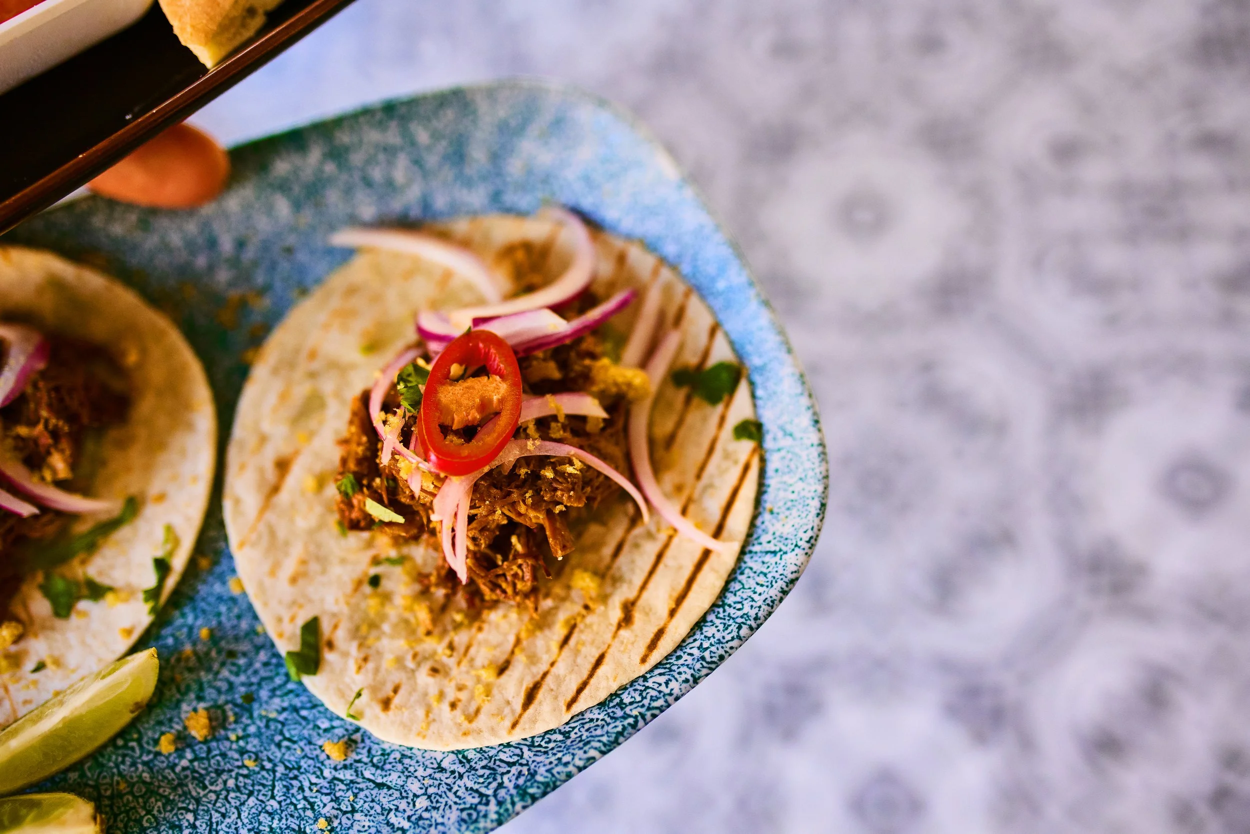 Close-up of two tacos with shredded meat, sliced red onions, and red pepper slices on a blue ceramic plate with lime wedges, on a gray textured surface.