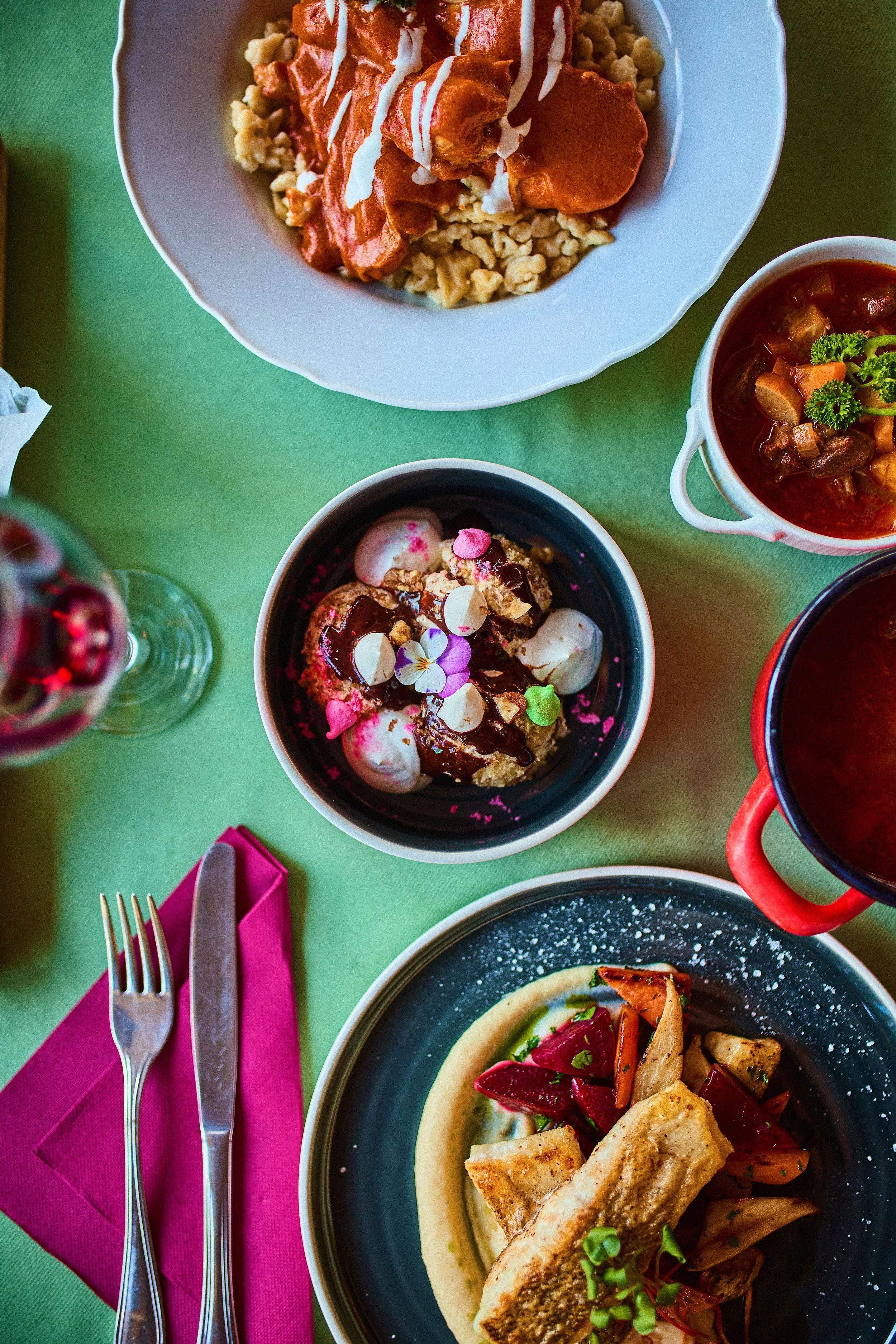 A top-down view of a table with various dishes, including a bowl of rice with chicken in red sauce with white drizzle, a bowl of dessert with marshmallows, chocolate syrup, and edible flowers, a bowl of beef stew, a plate of fish with vegetables on a