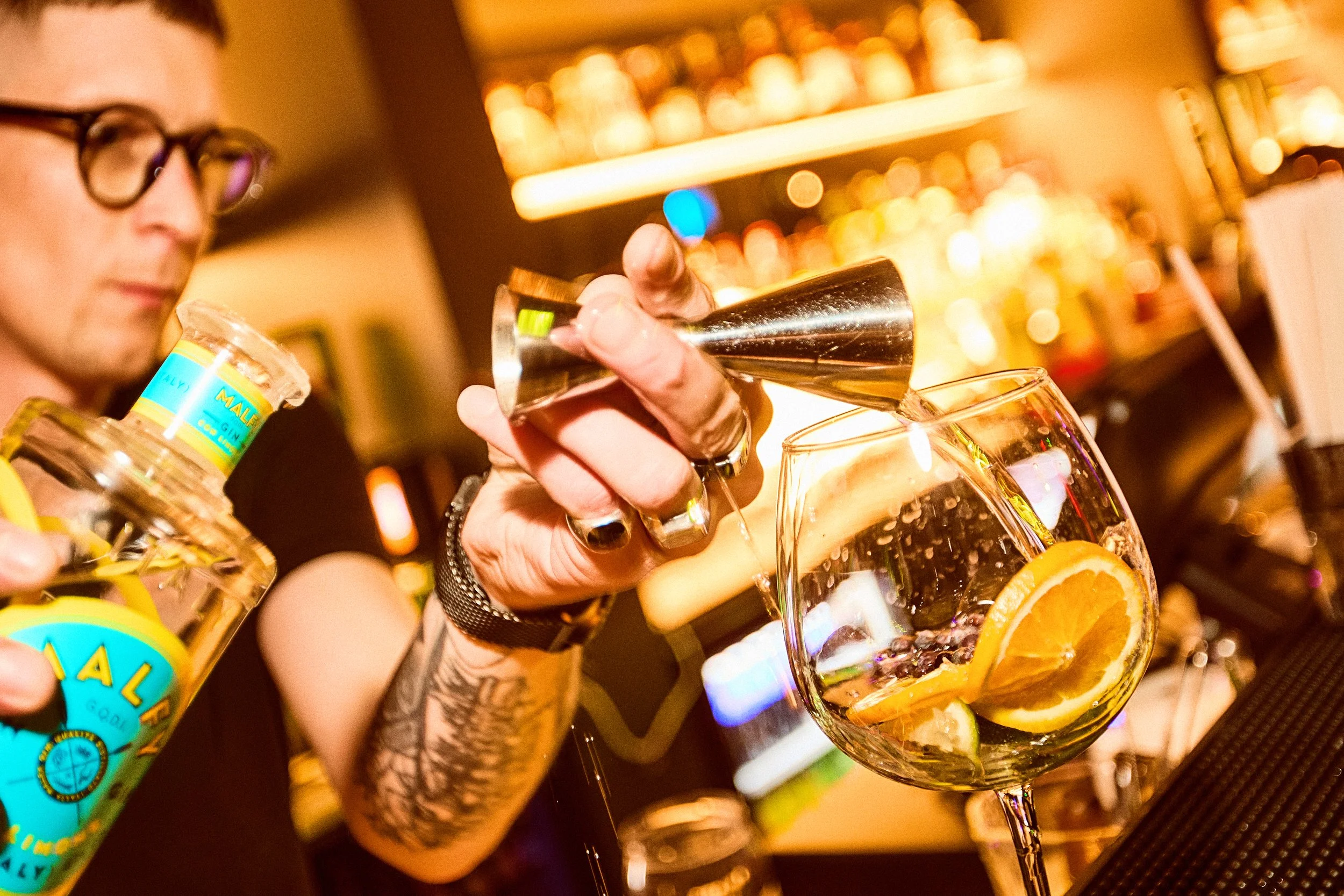 A bartender pours a clear liquid into a glass with orange slices at a bar, with blurred bottles in the background.