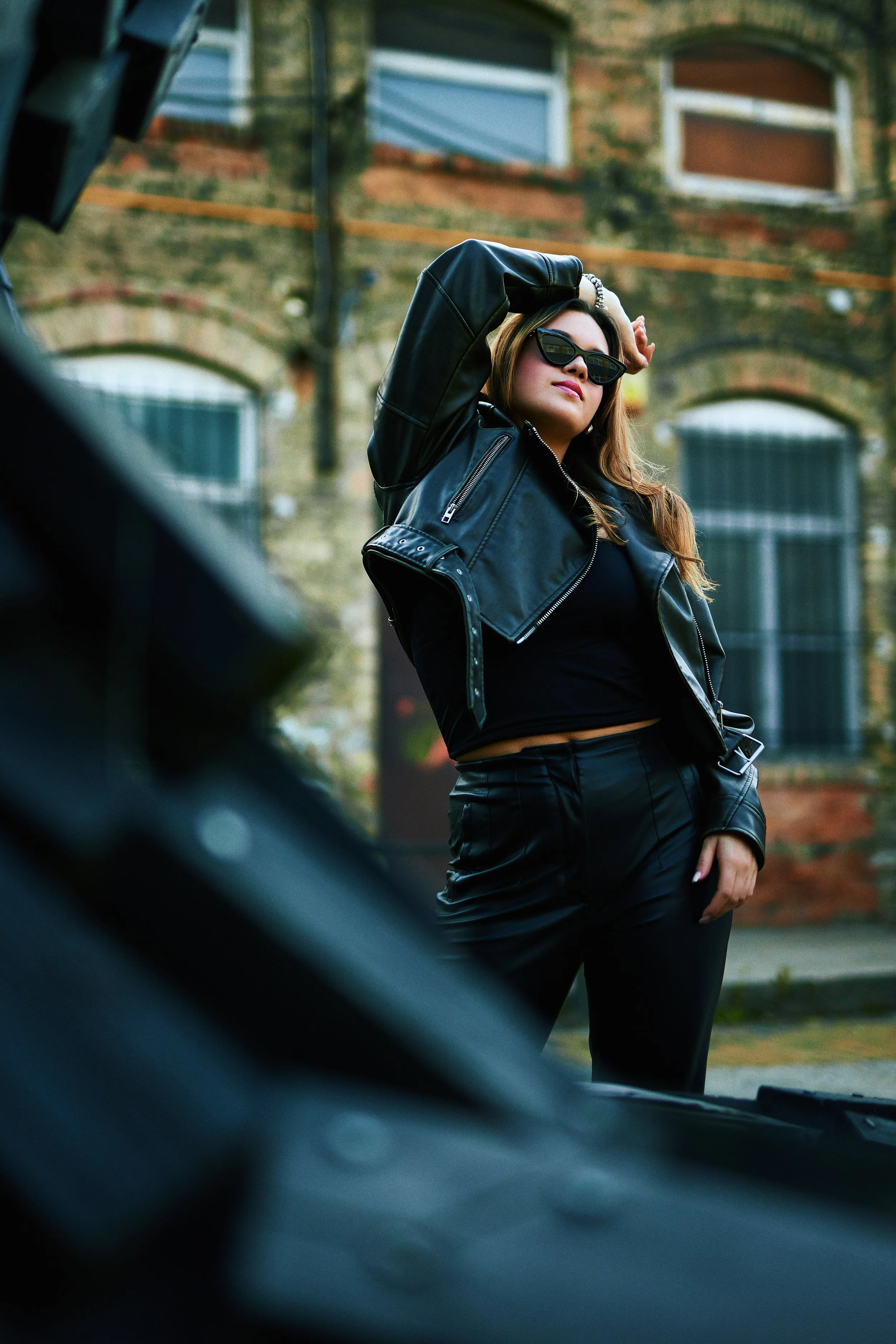 A woman with long hair wearing sunglasses, a black crop top, a black leather jacket, and black pants, posing with her arm raised behind her head in front of an old brick building.