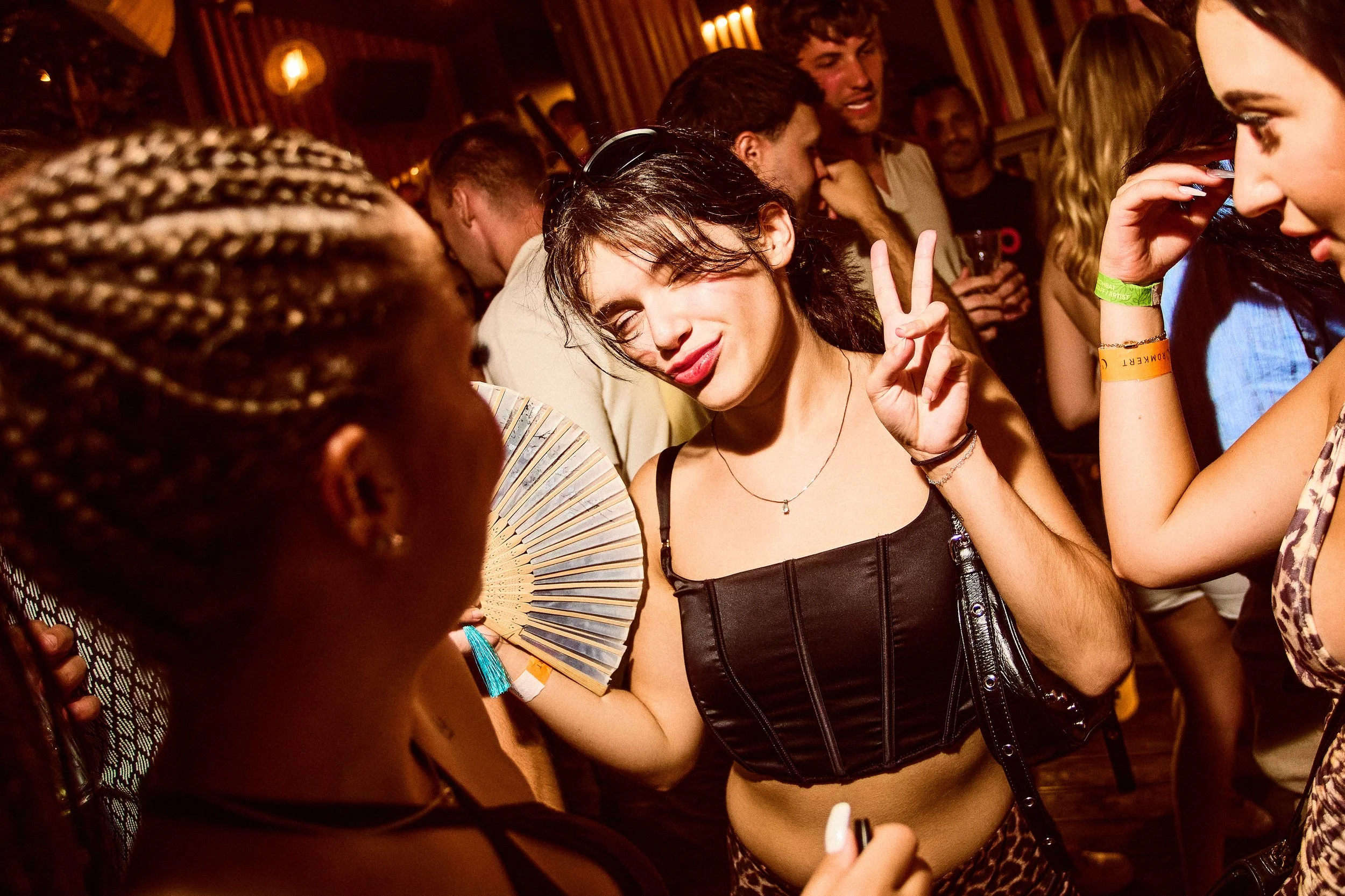 A group of young people dancing and socializing at a lively party or club, with a woman making a peace sign and smiling in the center