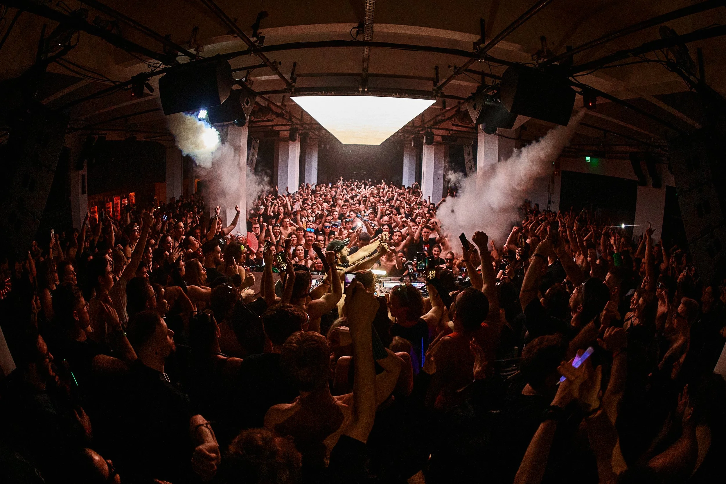 Crowd of people dancing and enjoying a DJ performance at a nightclub with smoke effects and large screen overhead.