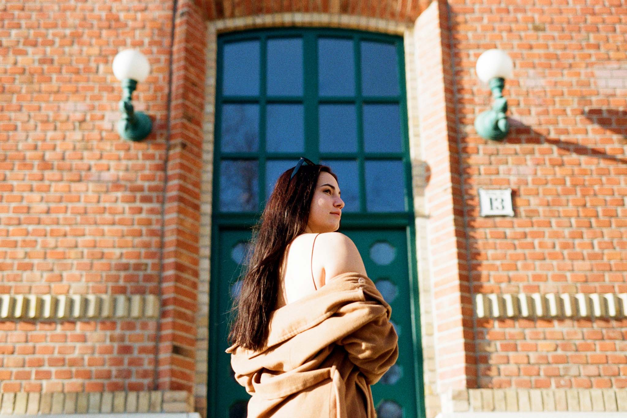 Young woman with long brown hair standing outside in front of a brick building with a green door and wall-mounted lamps, wearing a beige coat and glasses on her head.