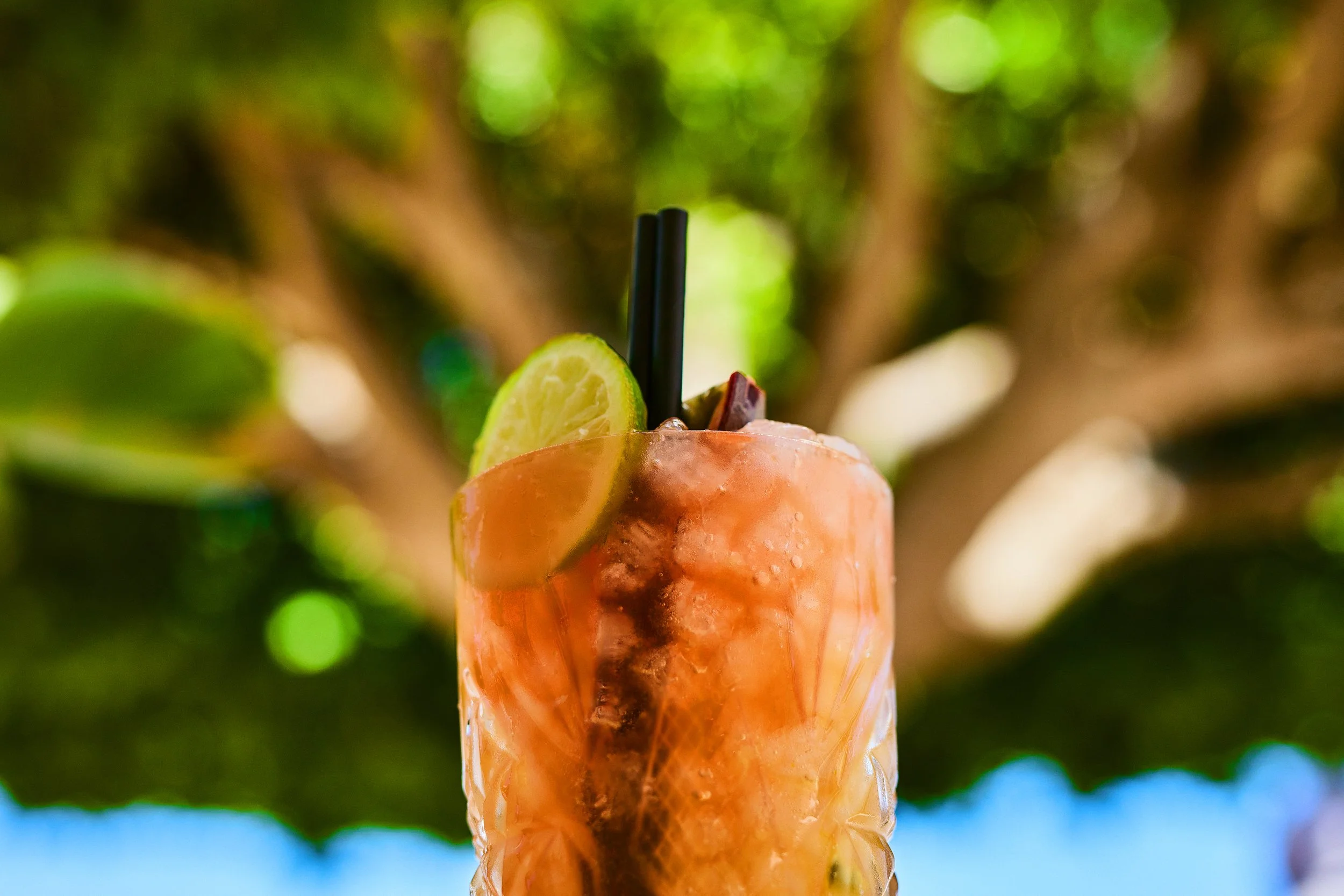 Close-up of a tropical cocktail with ice, garnished with a lime slice and a cherry, with a blurred green outdoor background.