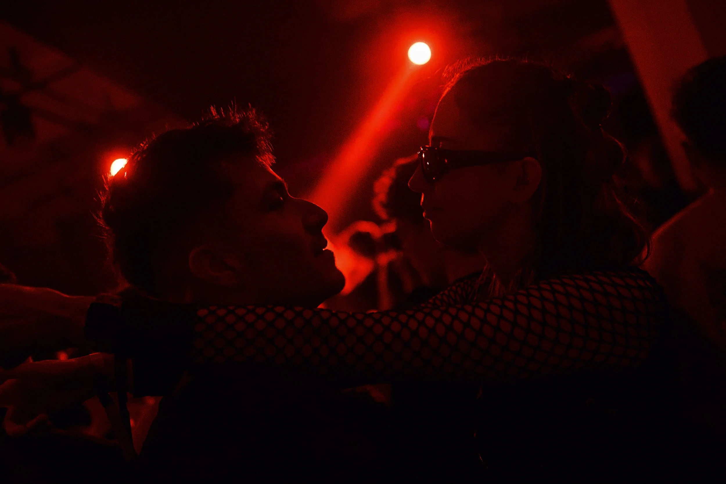 A couple dancing closely in a dimly lit setting with red lighting, silhouetted against the dark background.