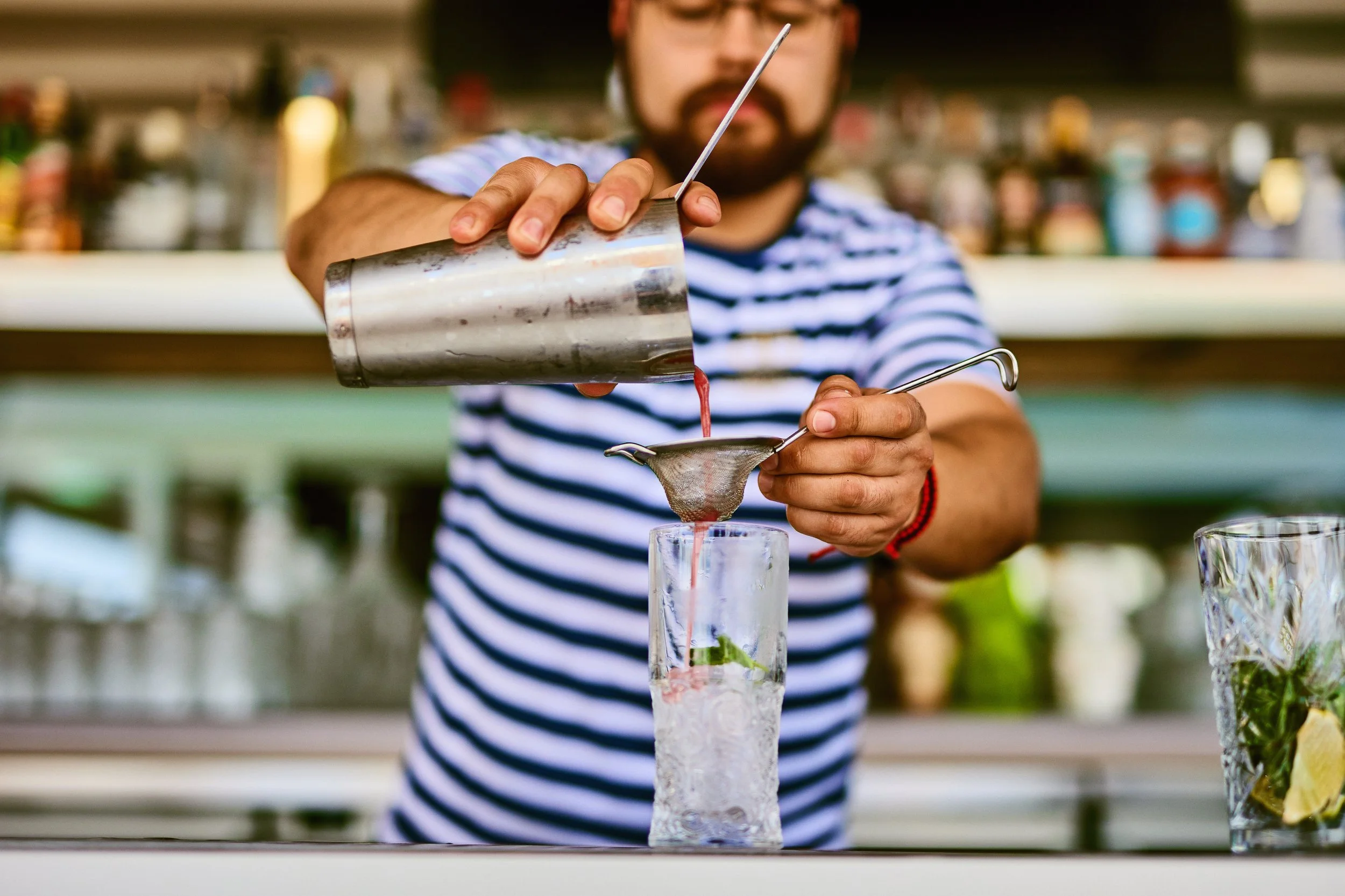 A bartender pouring a red cocktail through a strainer into a glass with ice and mint leaves.