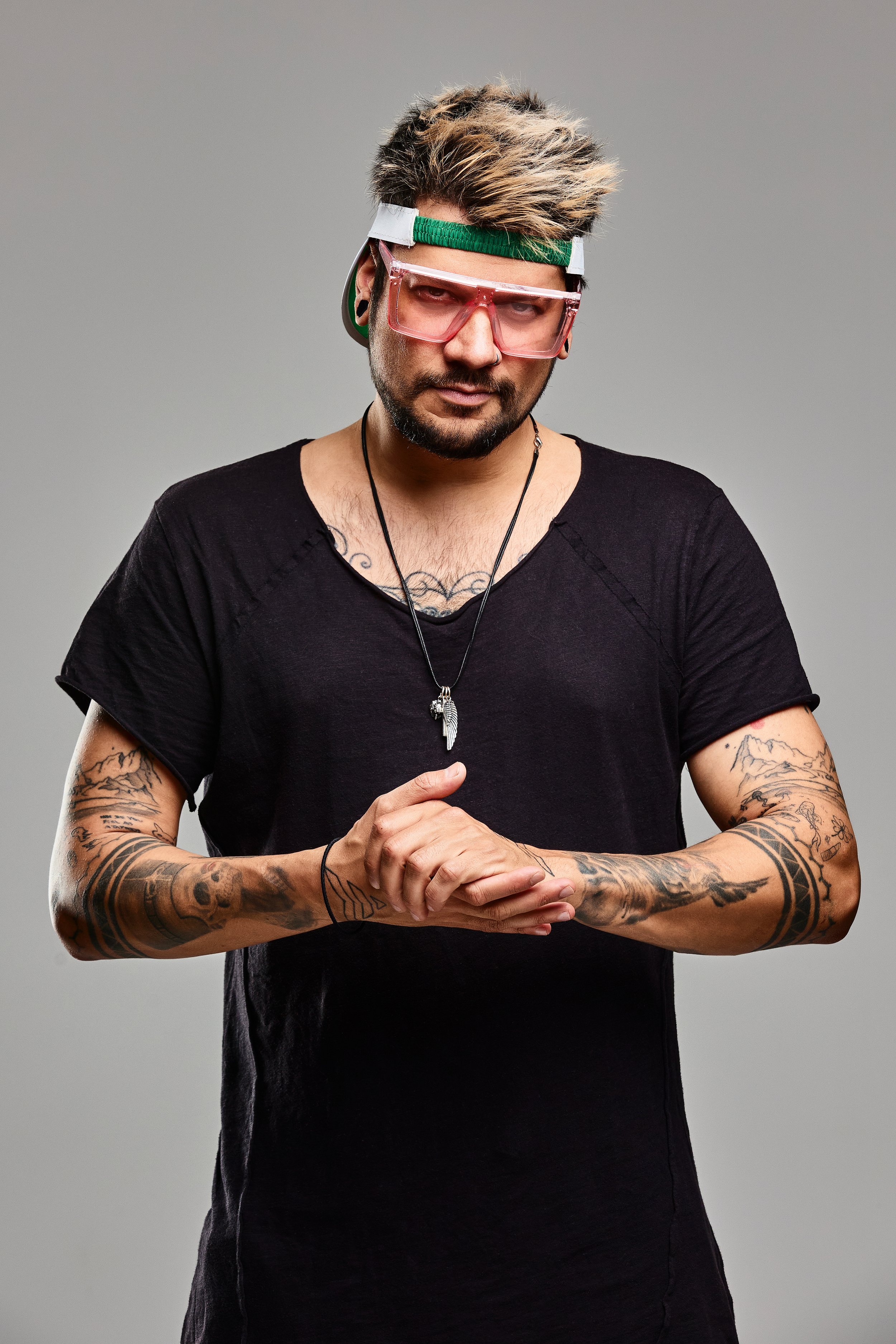 A man with styled, light-colored hair, wearing large pink sunglasses, a backwards green and white cap, and a black T-shirt. He has tattoos on his arms and is holding his hands together, looking directly at the camera.