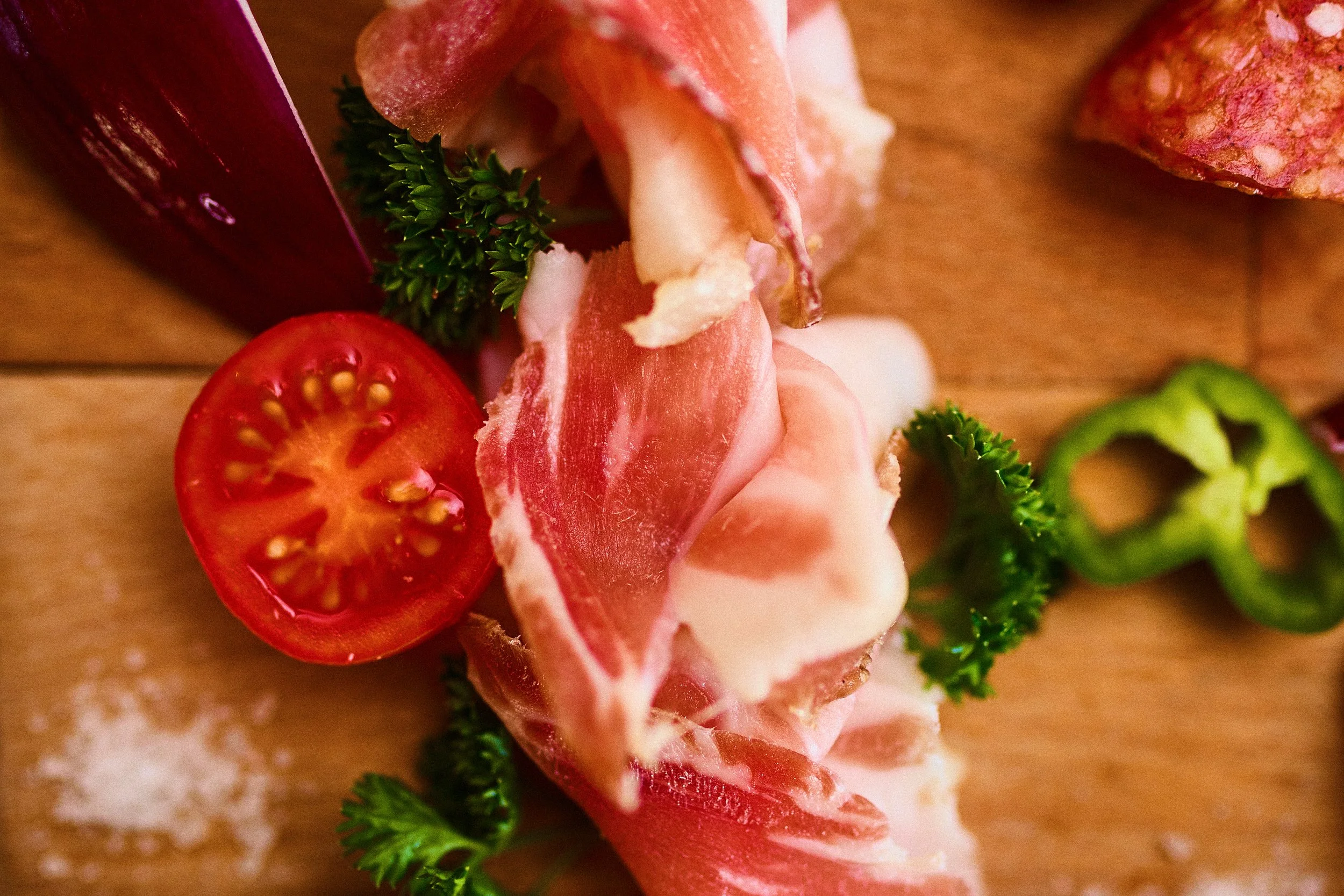 Close-up of cherry tomato, slices of cured meat, parsley, green pepper, and salt on a wooden surface.