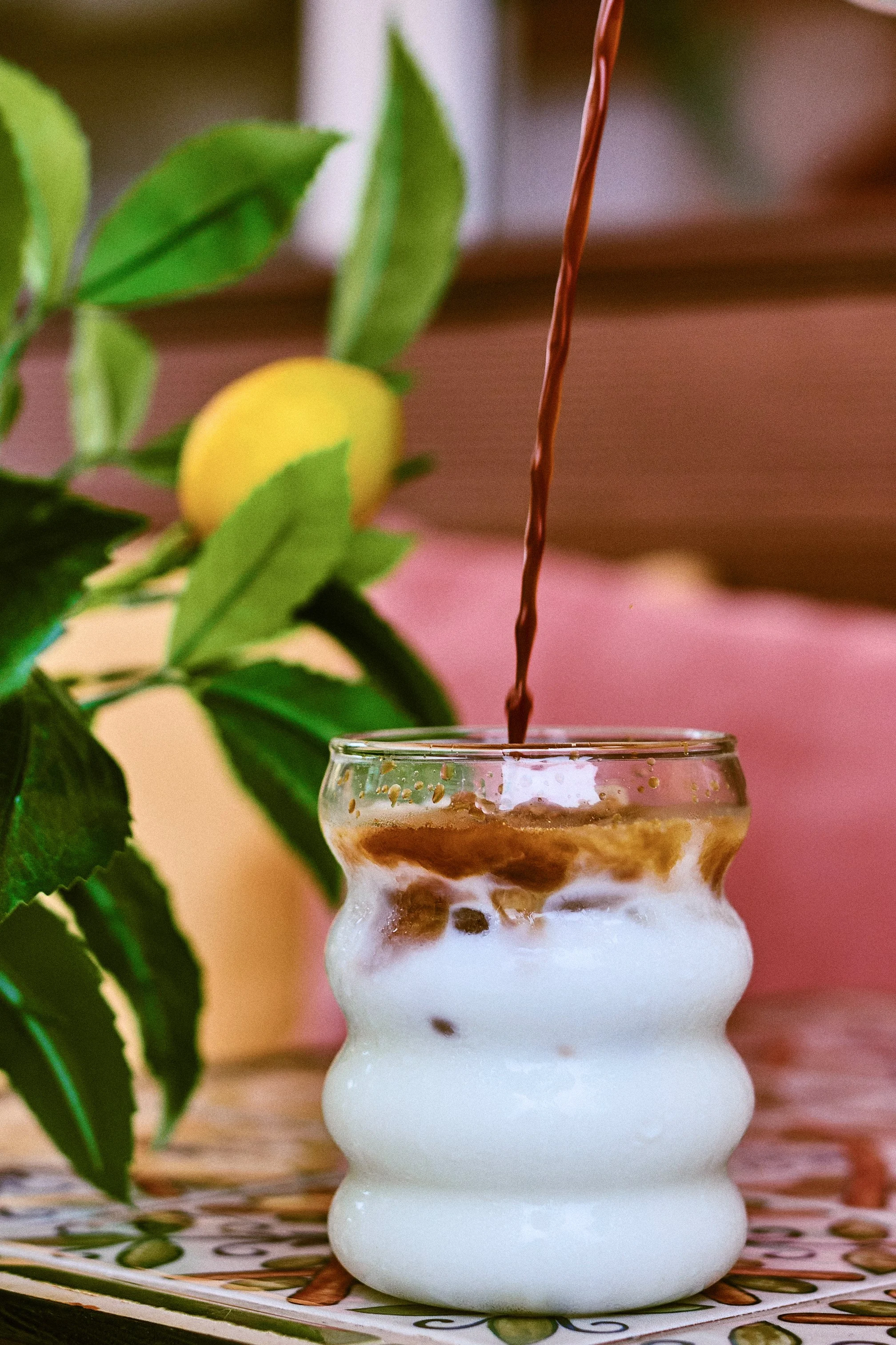 A glass with a wavy shape is filled with a layered drink, and a stream of chocolate syrup is being poured into it. There is a lemon and green leaves in the background.