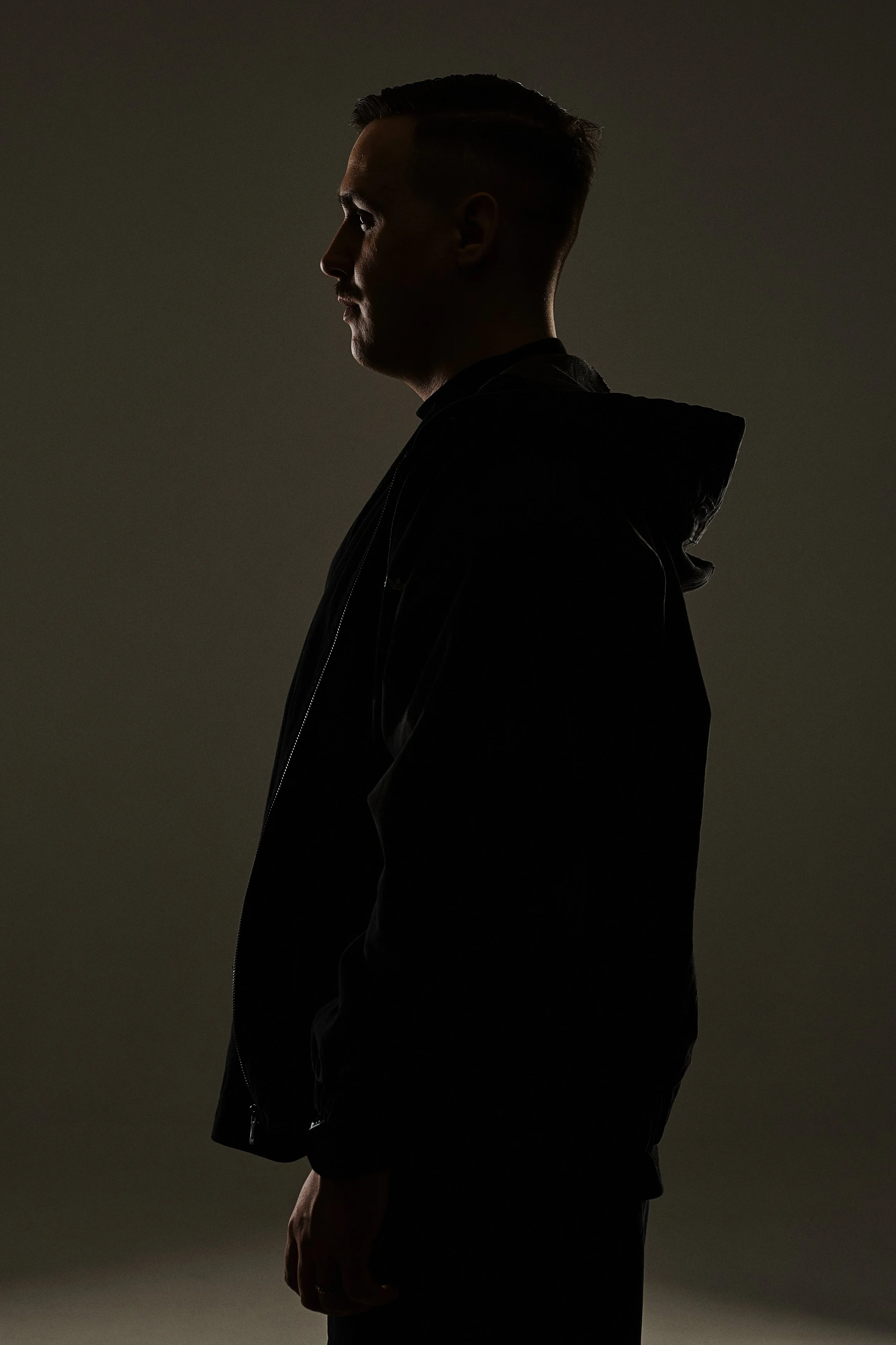 Profile silhouette of a young man with a fade haircut, wearing a black jacket, against a neutral background.