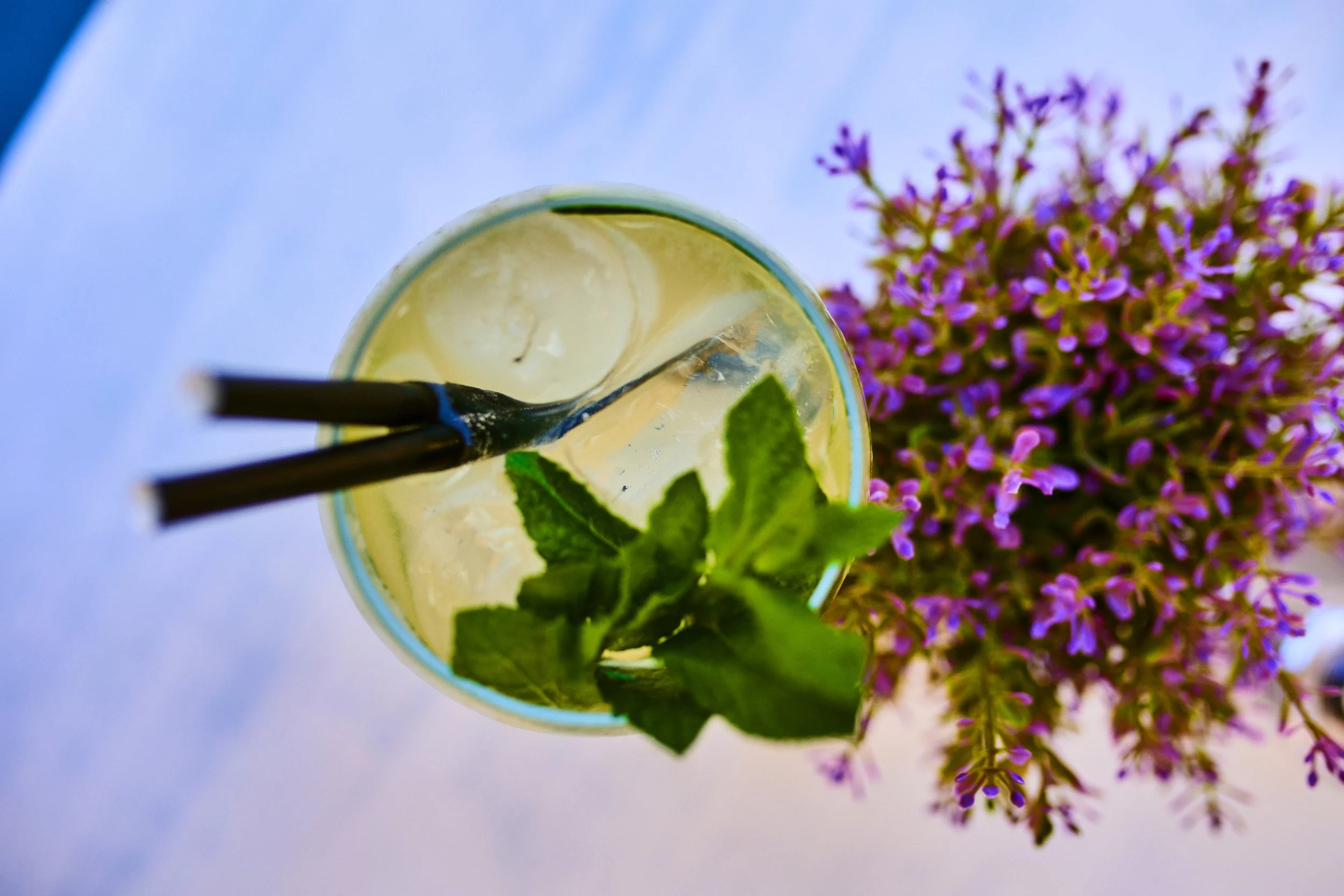 Top-down view of a glass with a yellow-green drink, ice cubes, black straws, and fresh mint leaves, next to a bunch of purple flowers on a white surface.