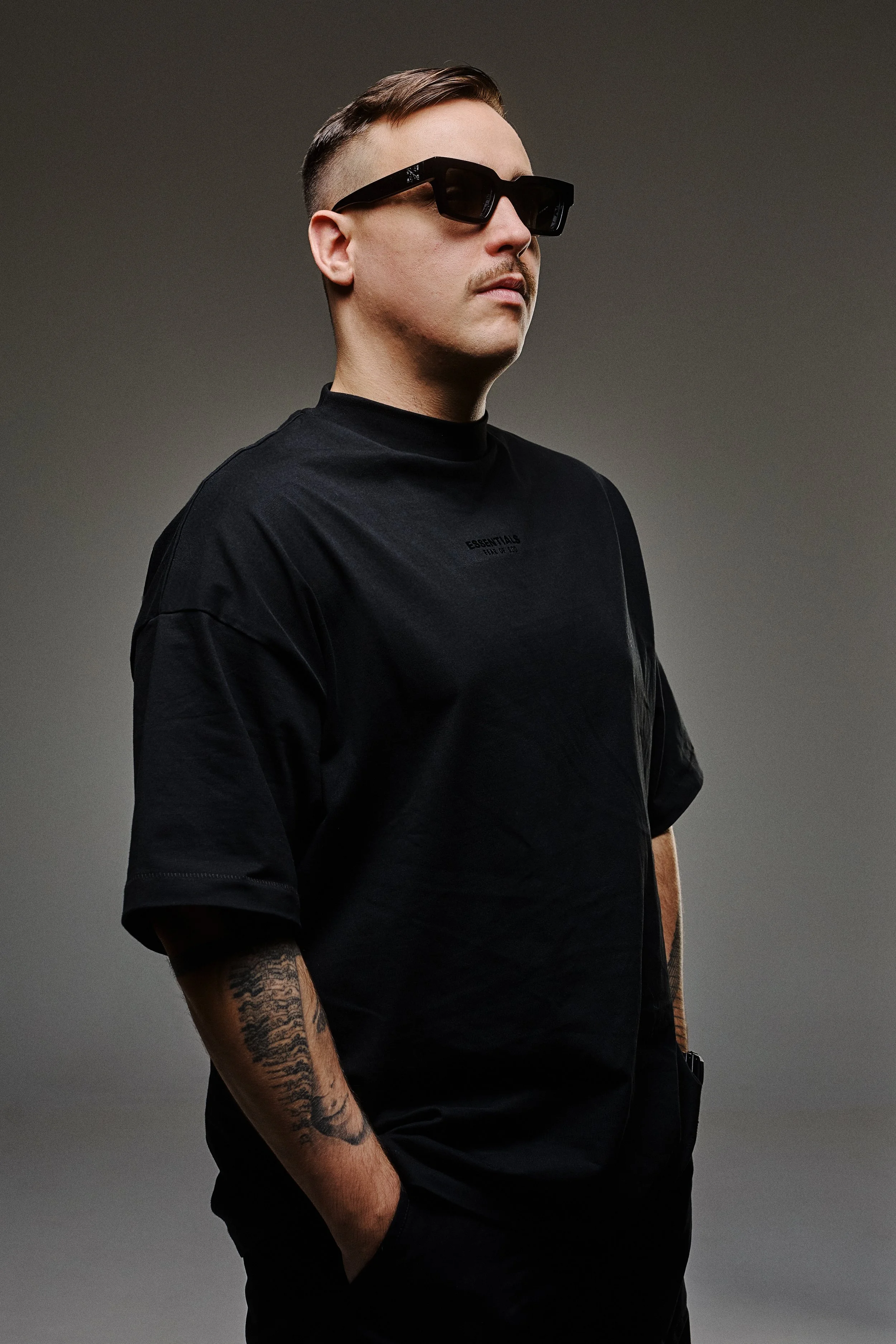 A man wearing black sunglasses and a black t-shirt with tattooed arms, standing against a gradient background.