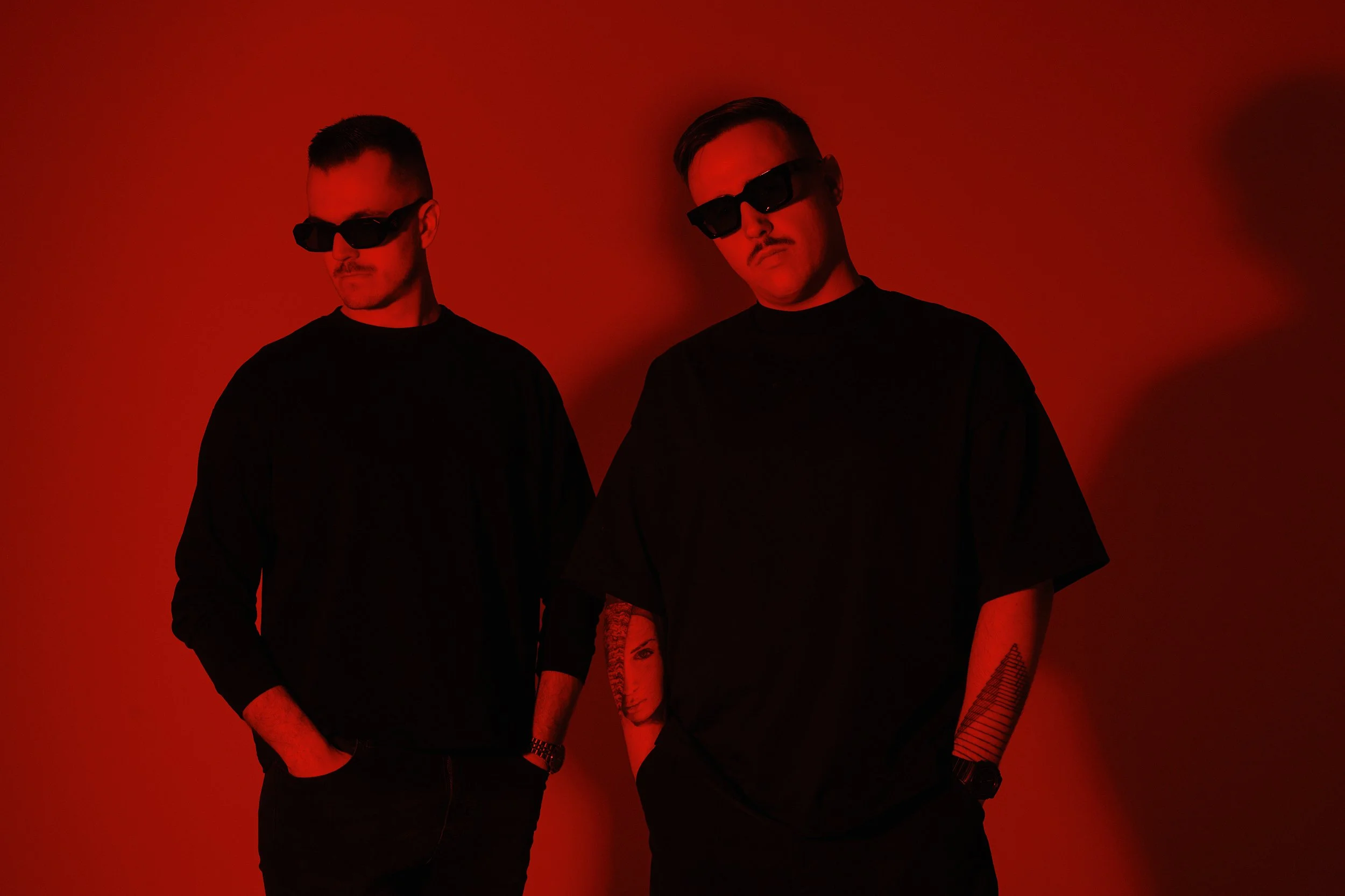 Two men wearing black sunglasses and black clothing pose against a red background. Both have tattoos on their arms.