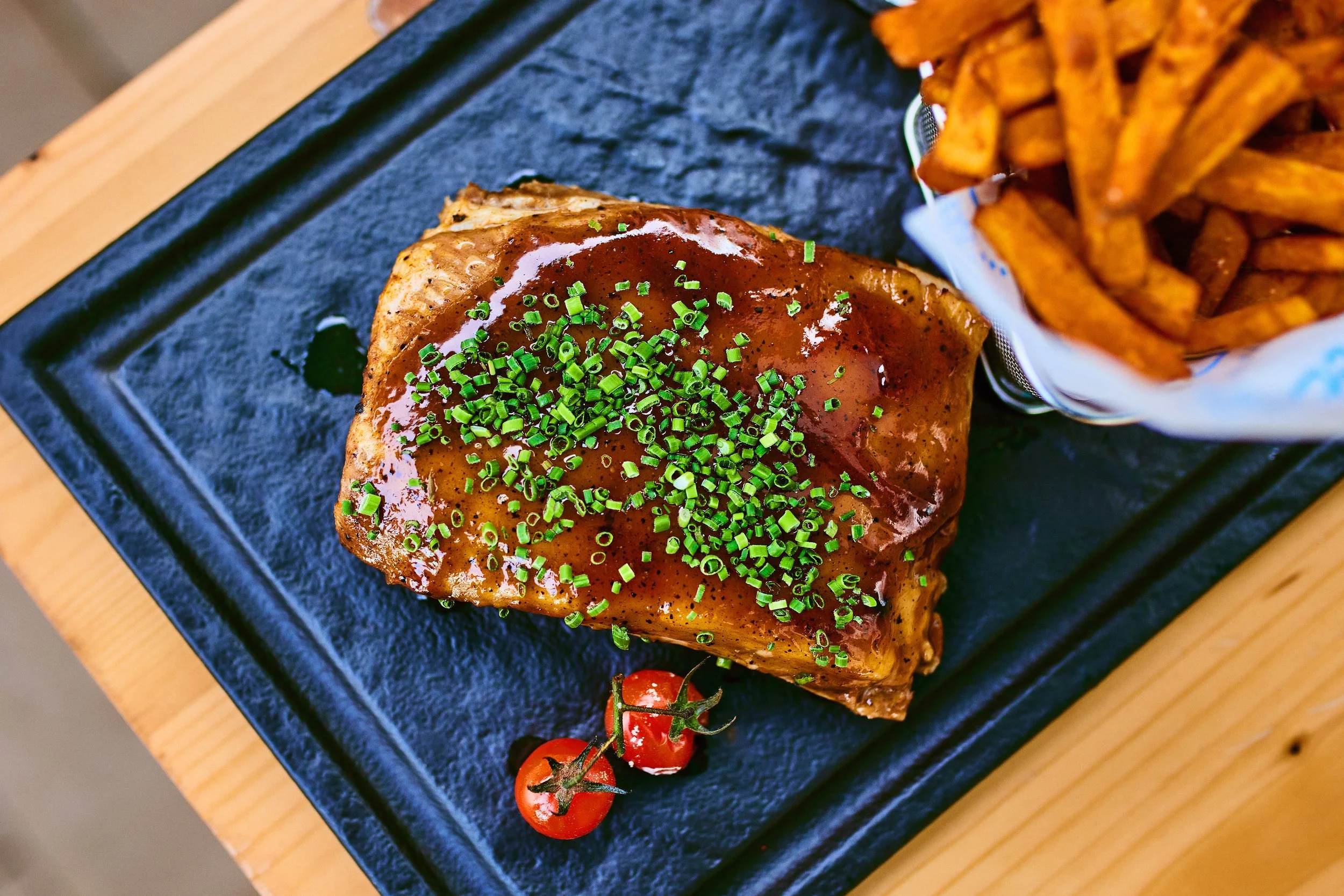 A serving of glazed meatloaf topped with chopped green onions, side of French fries in a paper container, and vine-ripened cherry tomatoes on a black slate serving platter on a wooden table.